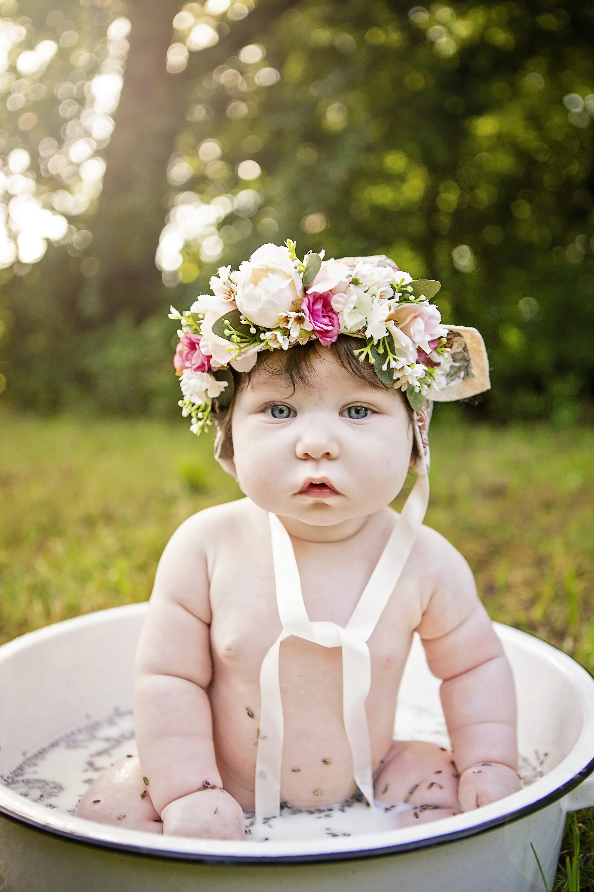 Baby sitting in a white basin filled with water and small bugs, wearing a flower crown, outdoors with greenery and sunlight in the background.