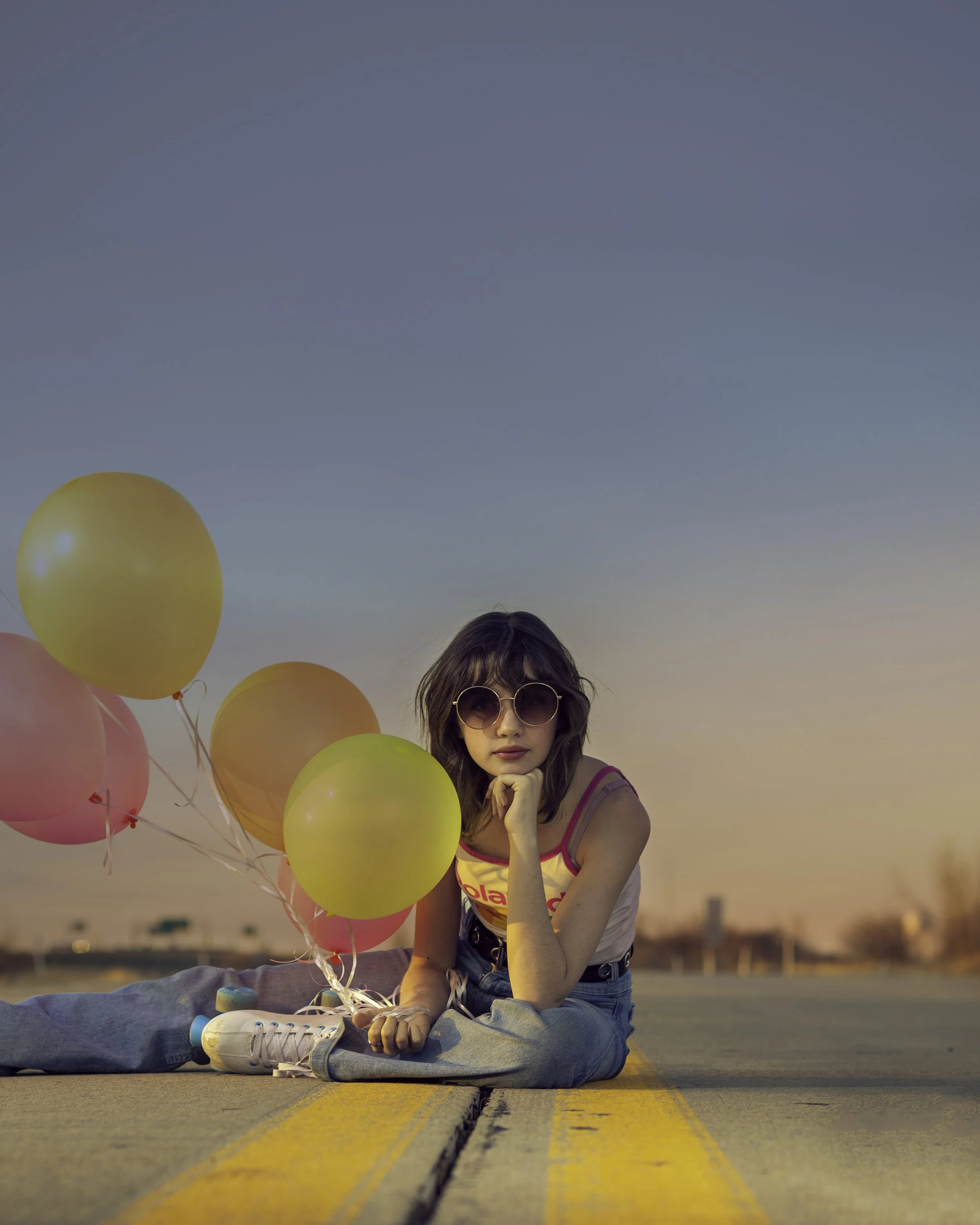 A young woman with sunglasses and wavy hair, sitting cross-legged on an empty road with a yellow line, holding pink and yellow balloons. The sky is clear with the setting sun in the background.