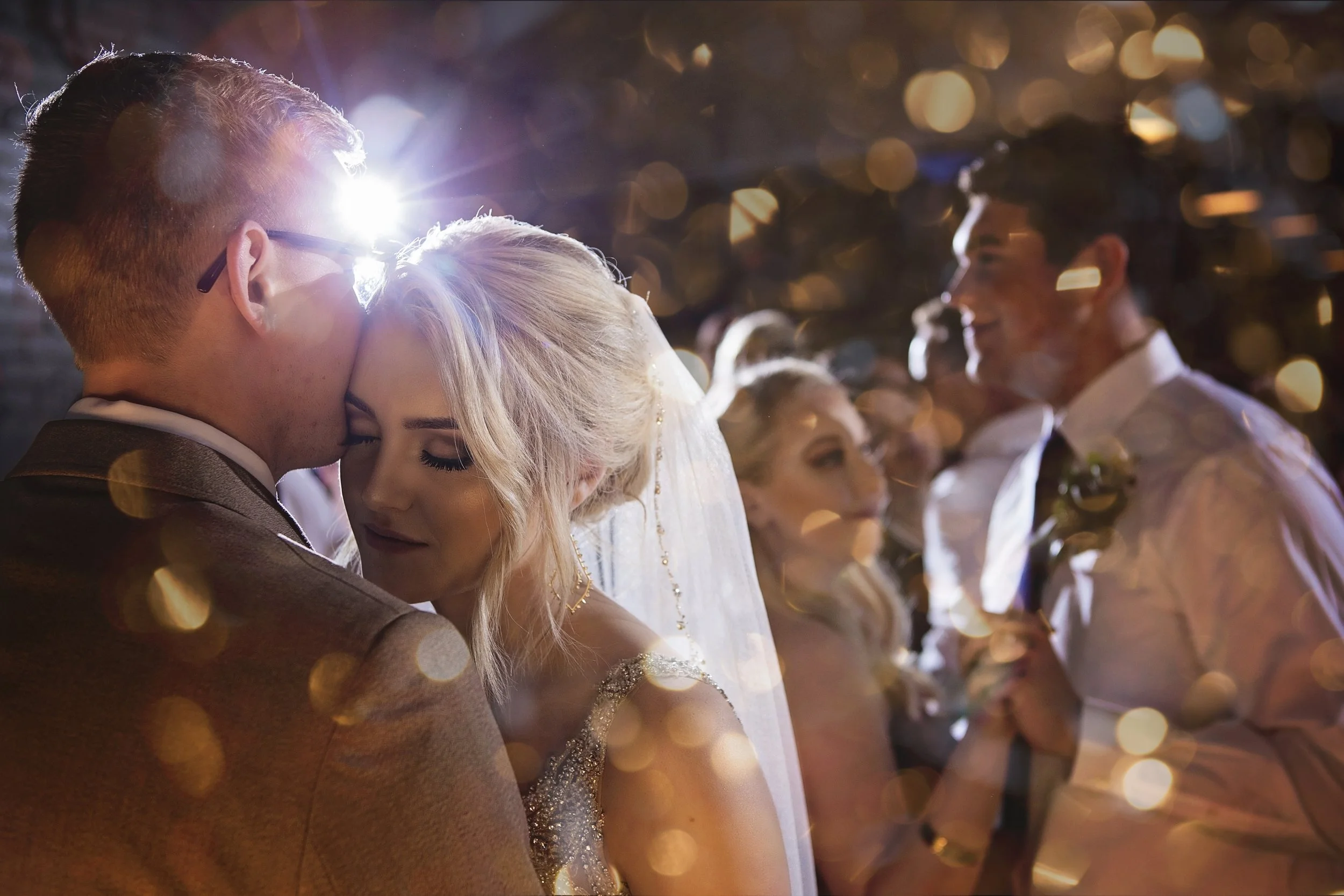 A bride and groom embrace closely on their wedding night, with a groom and bride dancing behind them in a romantic setting with bokeh lights.