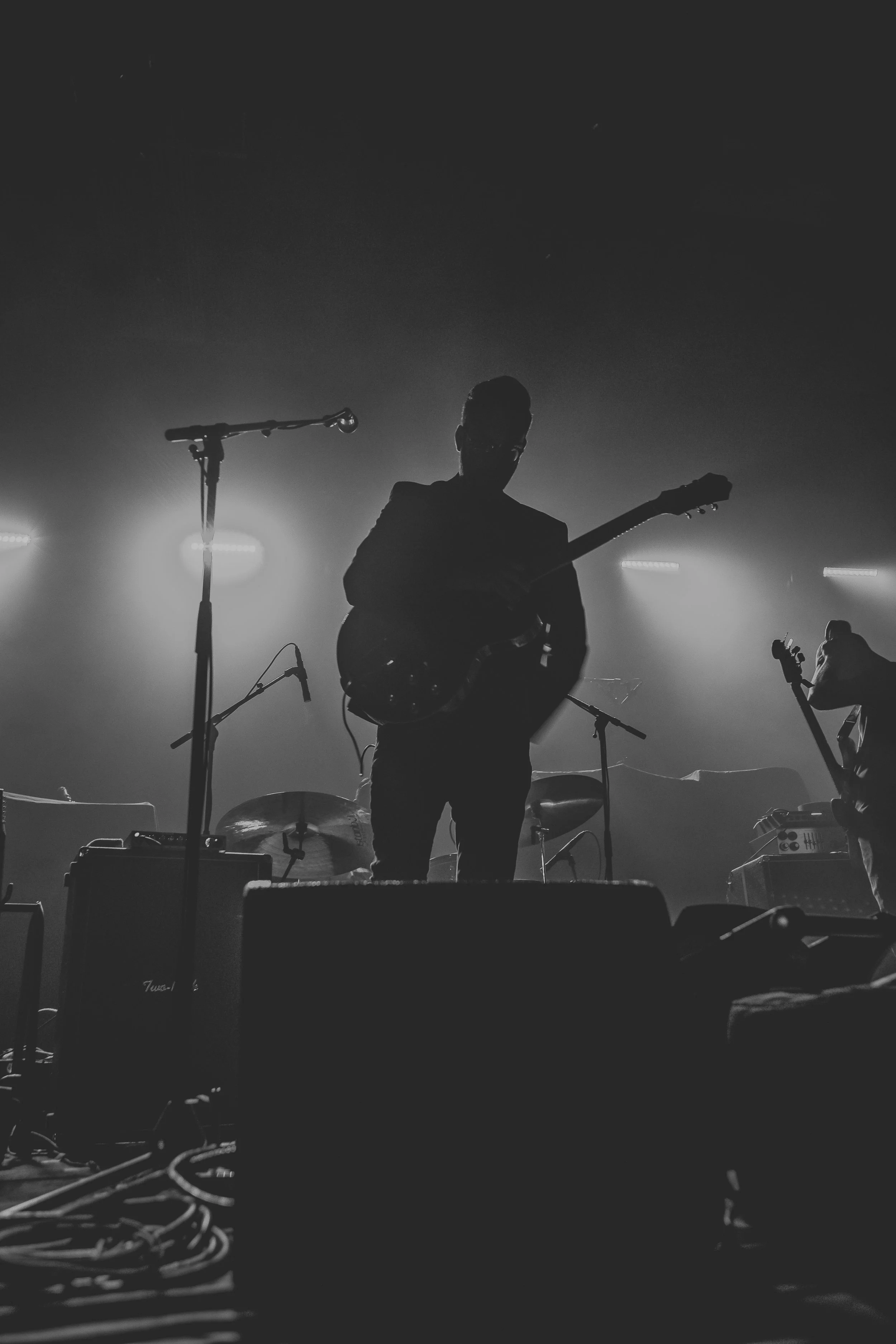 Silhouette of a musician with a guitar performing on stage under bright backlighting, with musical equipment and cables in the foreground, black and white photo.