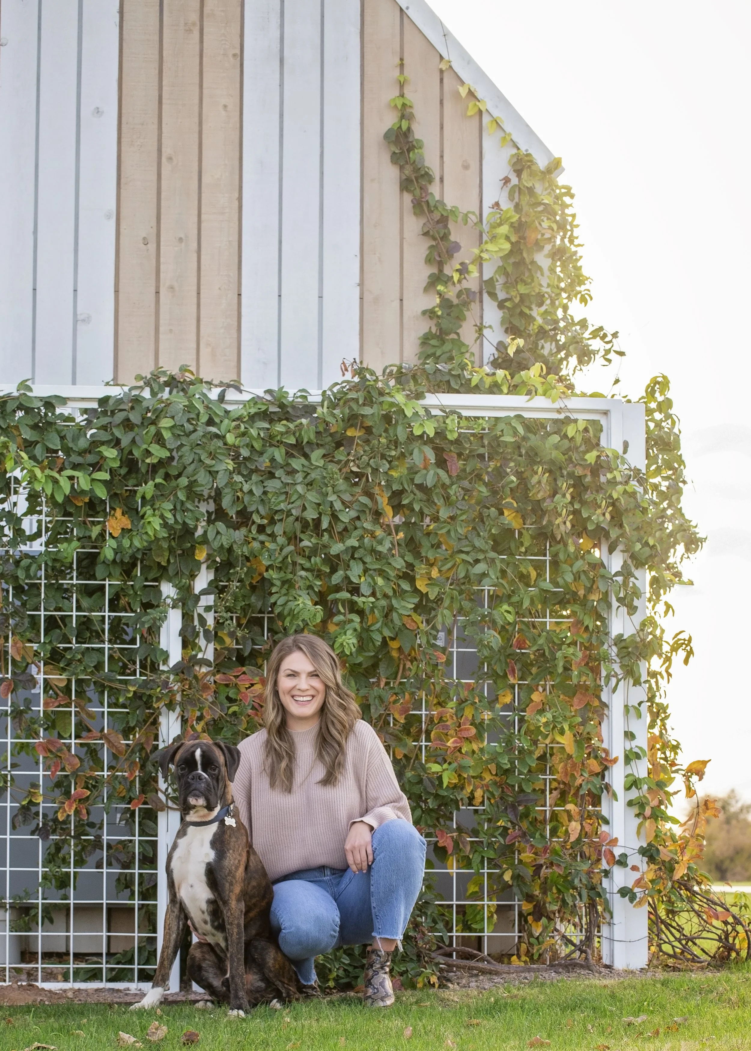 A woman smiling and sitting next to a large dog in front of a lush green vine-covered trellis outside a building with a wood and metal siding.