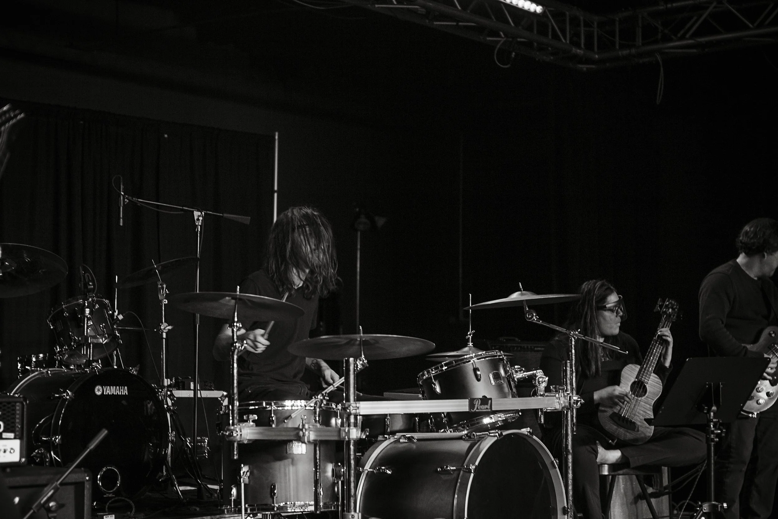 Black and white photo of a band performing on stage, with a drummer and a guitarist visible.