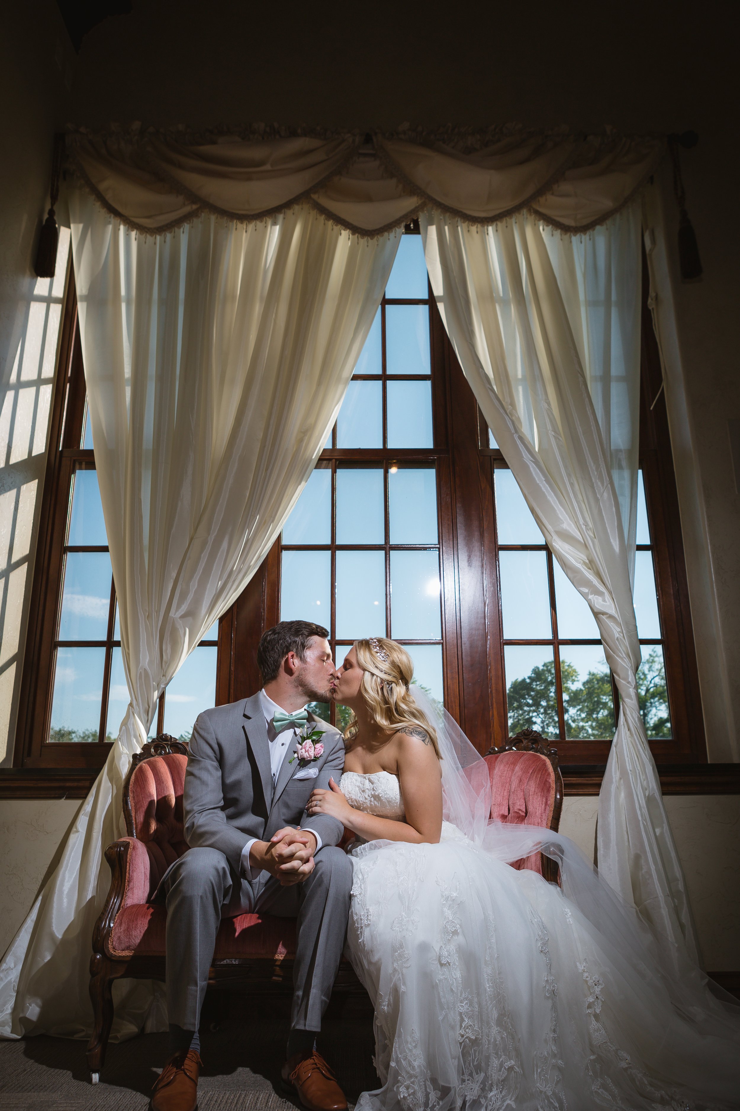 A bride and groom sitting on a vintage pink sofa, sharing a kiss inside a room with large windows and cream-colored curtains.