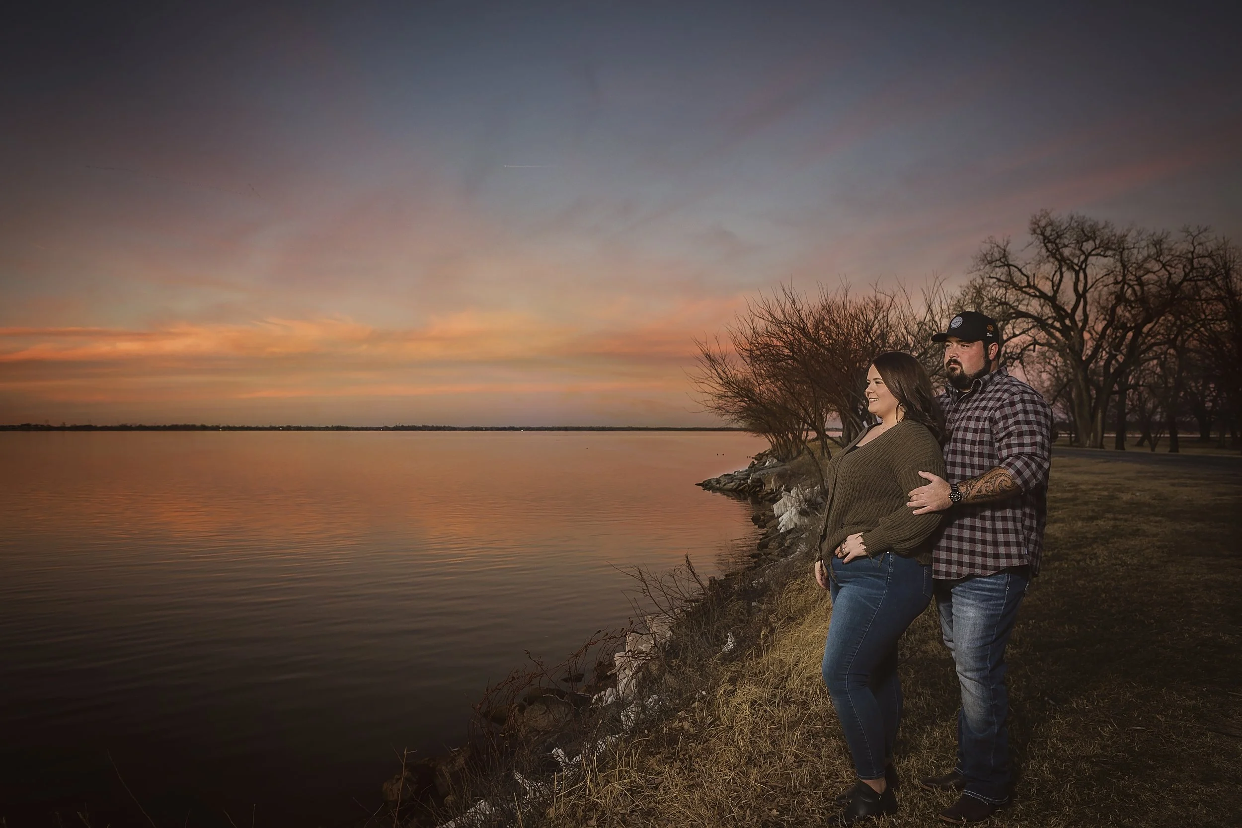 Engagement session by a lake beach at sunset, with a sky filled with pink and orange hues, and trees along the shoreline.