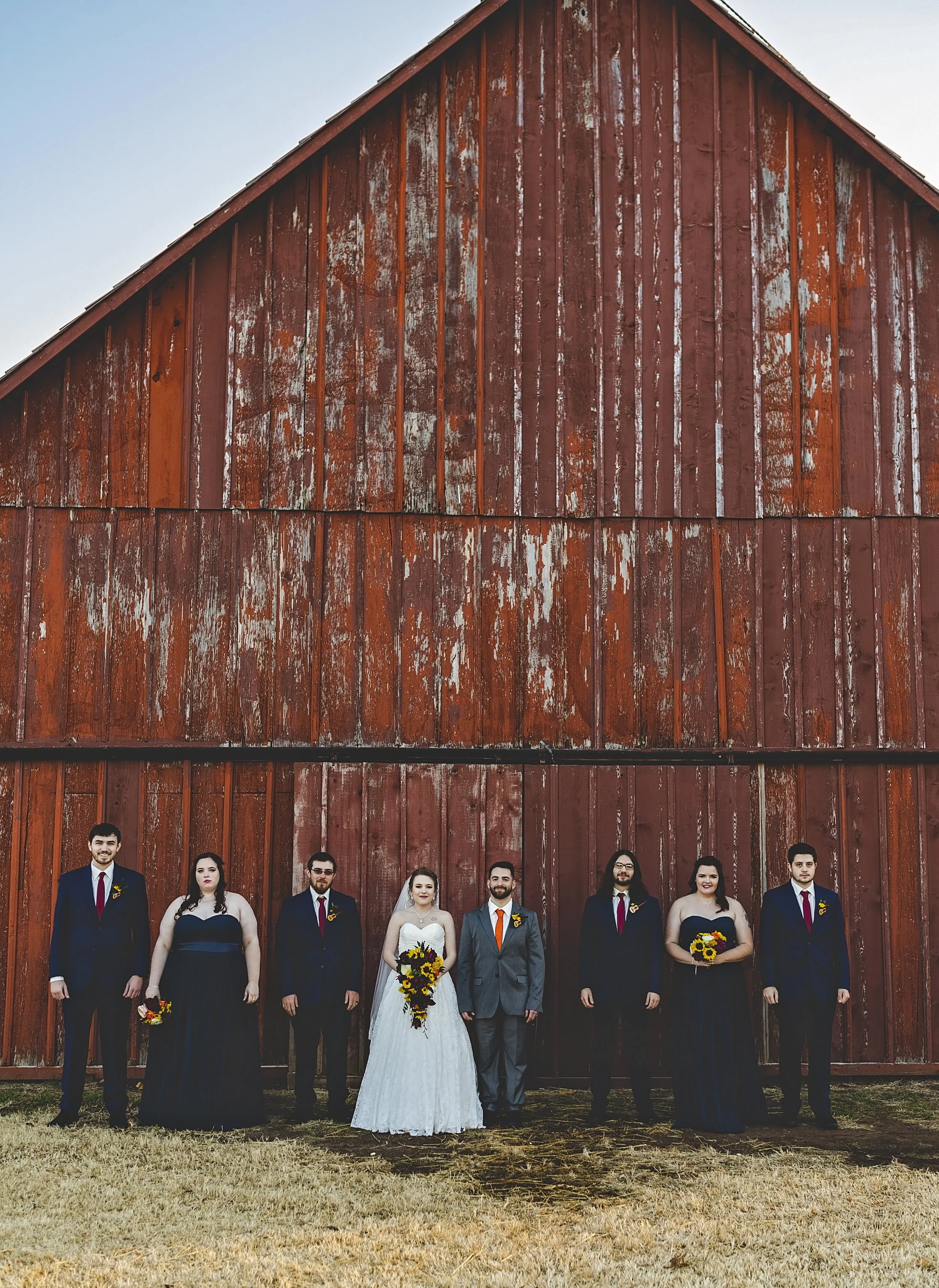 A wedding party of eight people standing in front of a large, weathered red barn.
