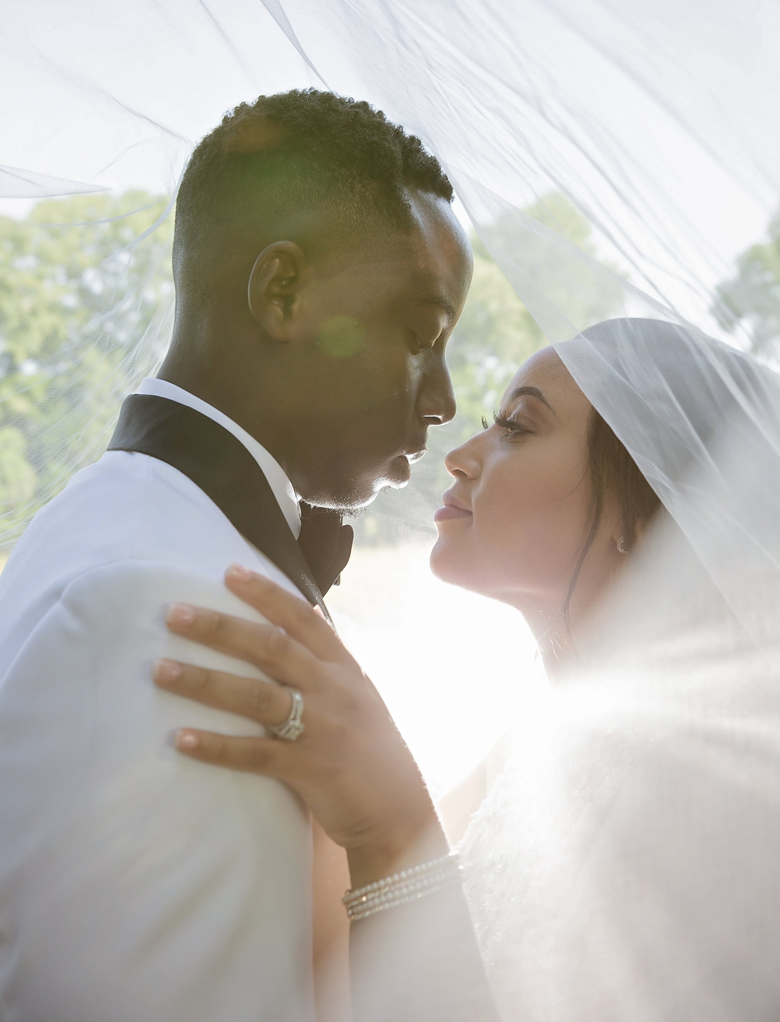 A bride and groom face each other closely under a wedding veil, with sunlight shining through, creating a romantic scene.