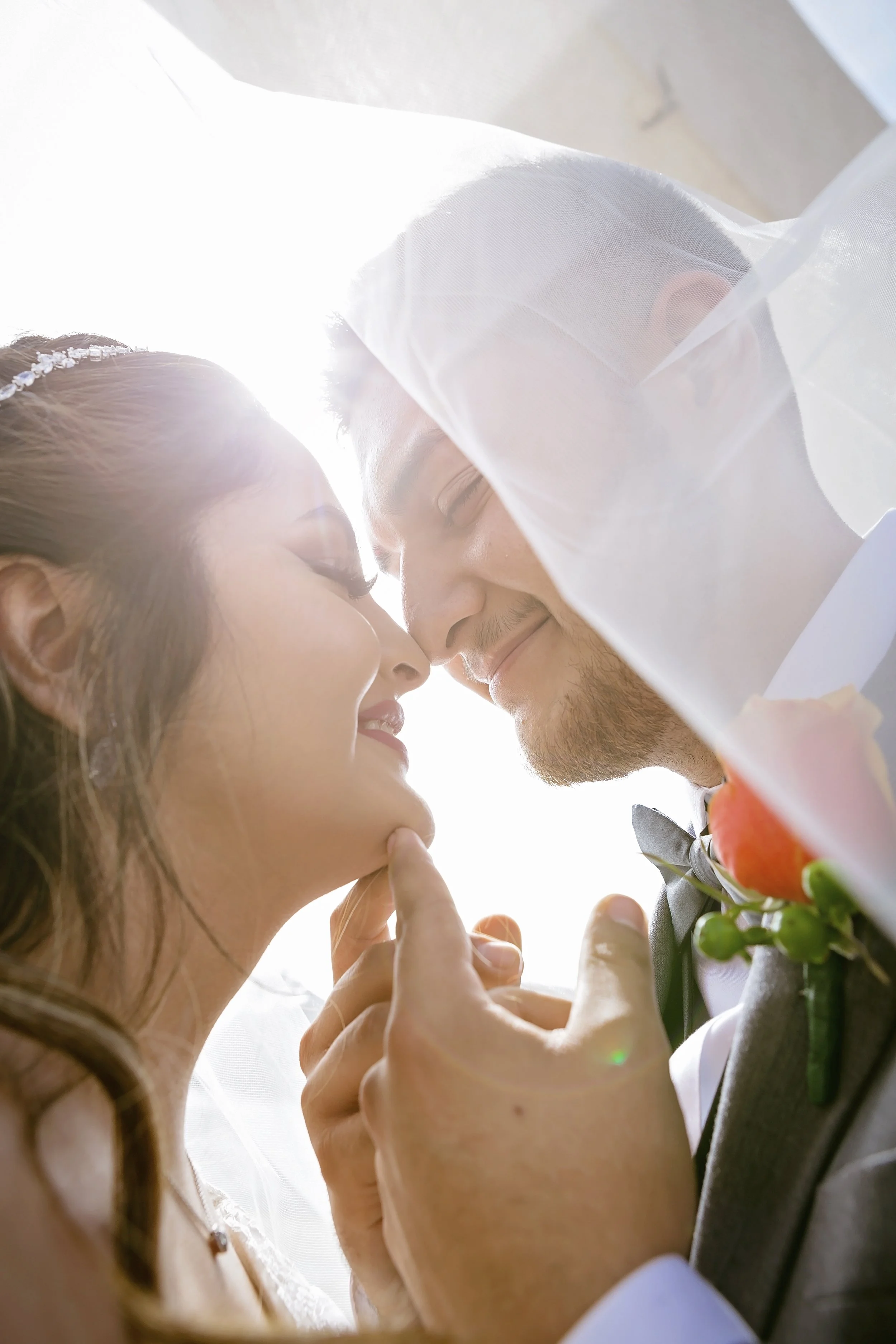 A close-up of a bride and groom touching foreheads with eyes closed, smiling, under a veil with bright backlight, wedding attire and floral boutonniere visible.