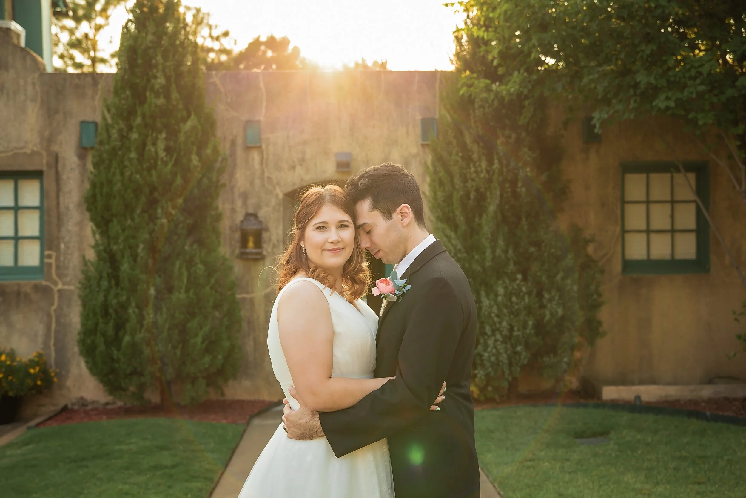 A couple in wedding attire, the bride in a white gown and the groom in a black suit, holding each other closely and smiling in a garden at sunset.
