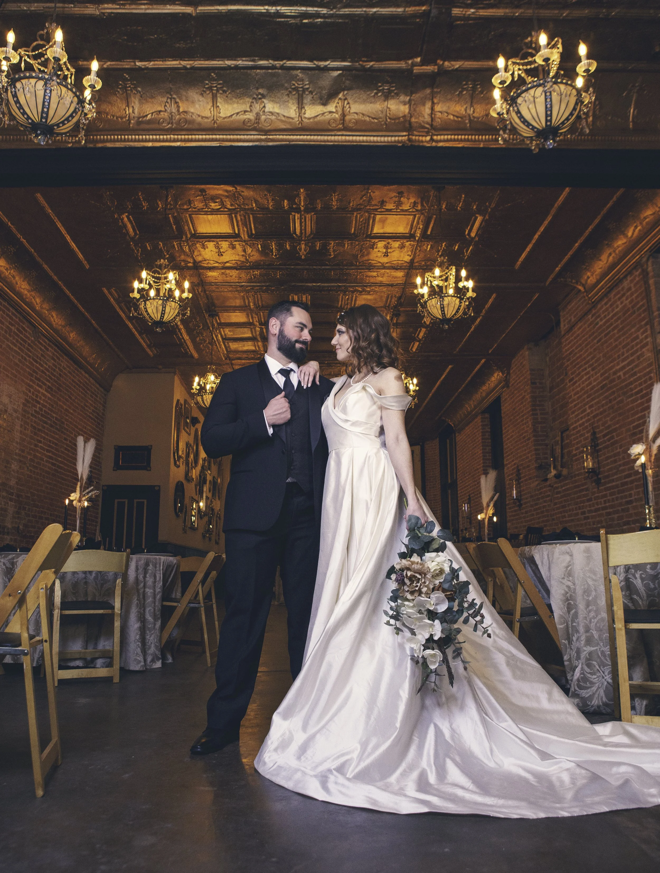 A bride and groom standing close together in a decorated wedding reception hall, with the bride holding a bouquet and the groom dressed in a suit. They are indoors with ornate ceiling chandeliers and brick walls.