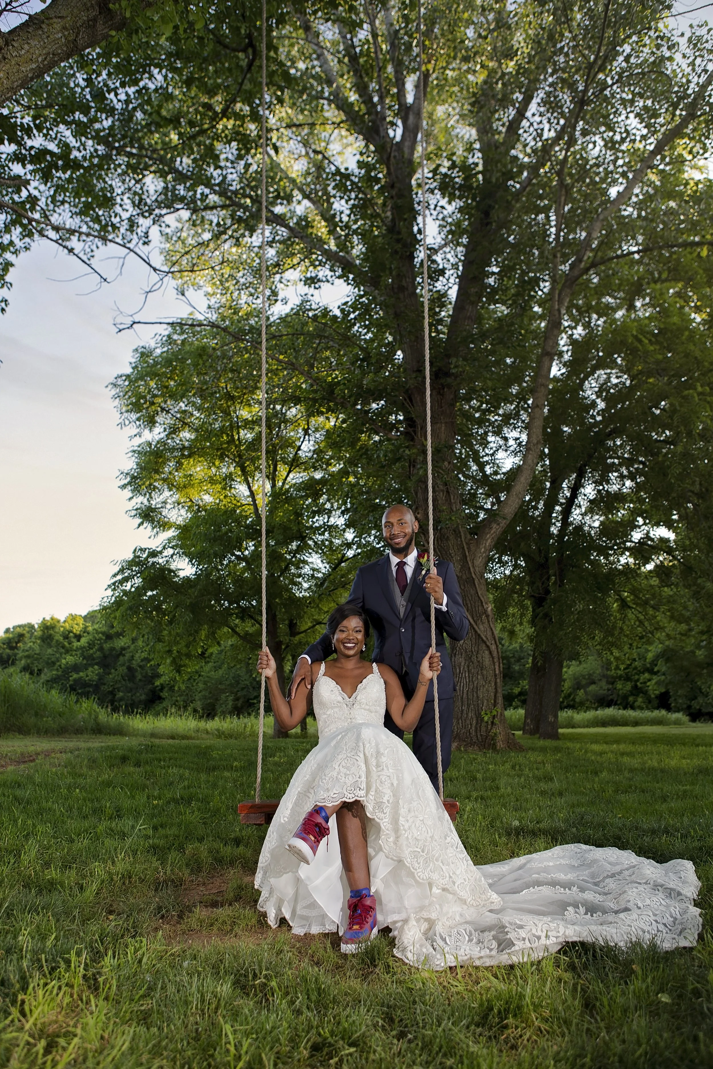 A bride in a white wedding gown sitting on a swing, with a groom standing behind her, outdoors under a large tree during daytime.