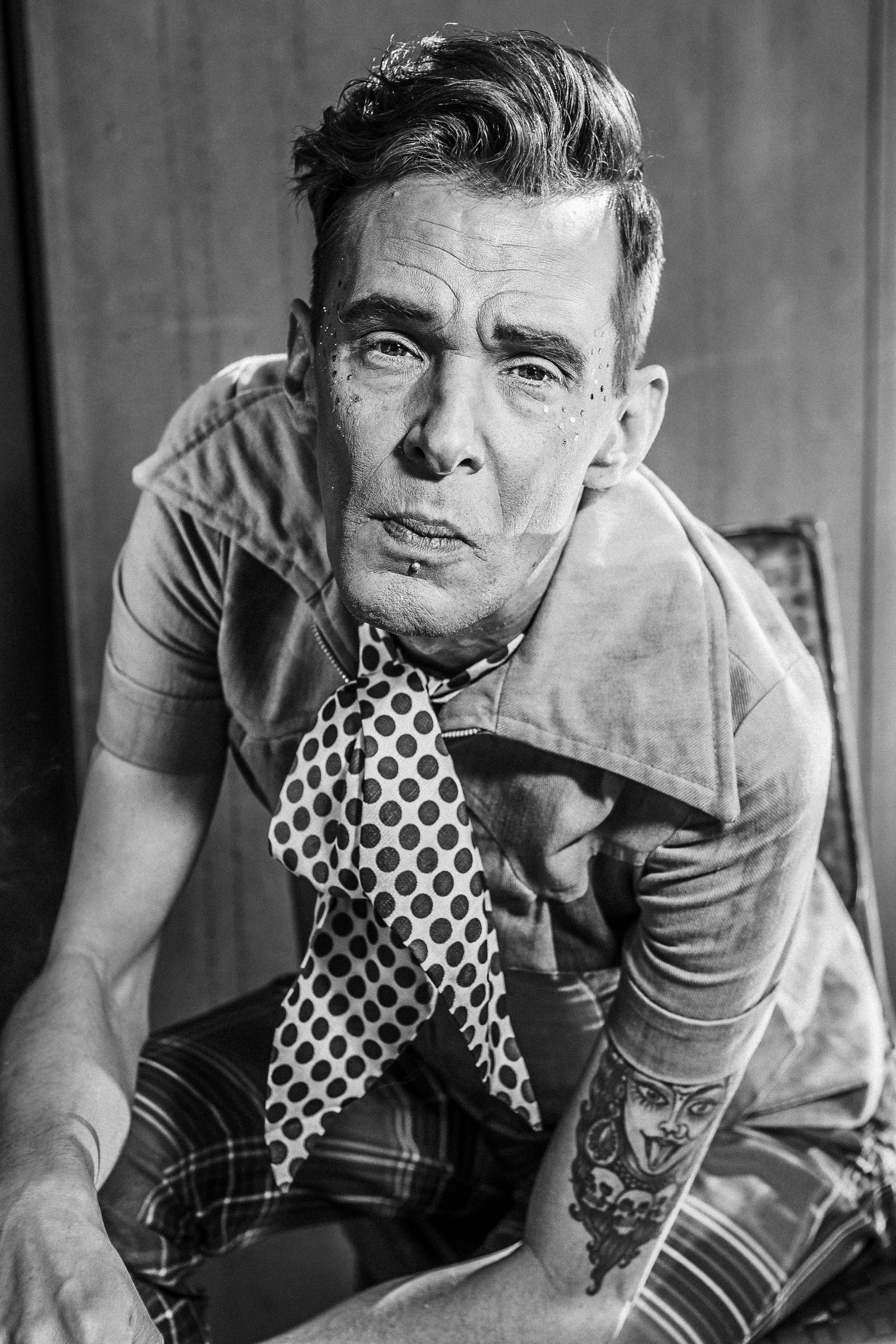 Black and white photo of a man with tattoos, wearing a patterned bandana and plaid pants, looking directly at the camera in a close-up shot.