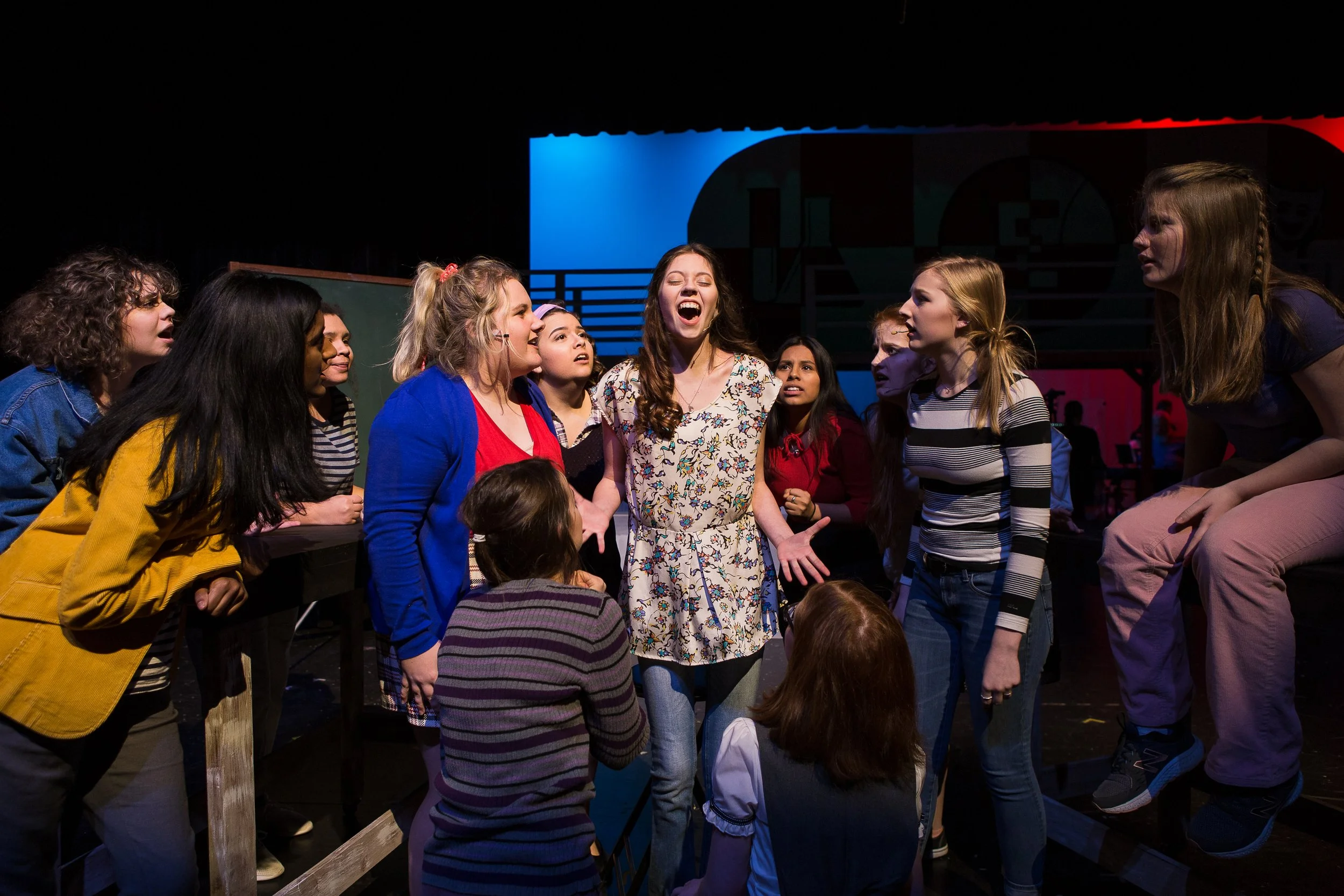 A group of young women acting in a theatrical rehearsal or performance. They are gathered around one woman who is speaking or singing passionately, with various facial expressions and engaging body language. The background suggests a stage setting wi