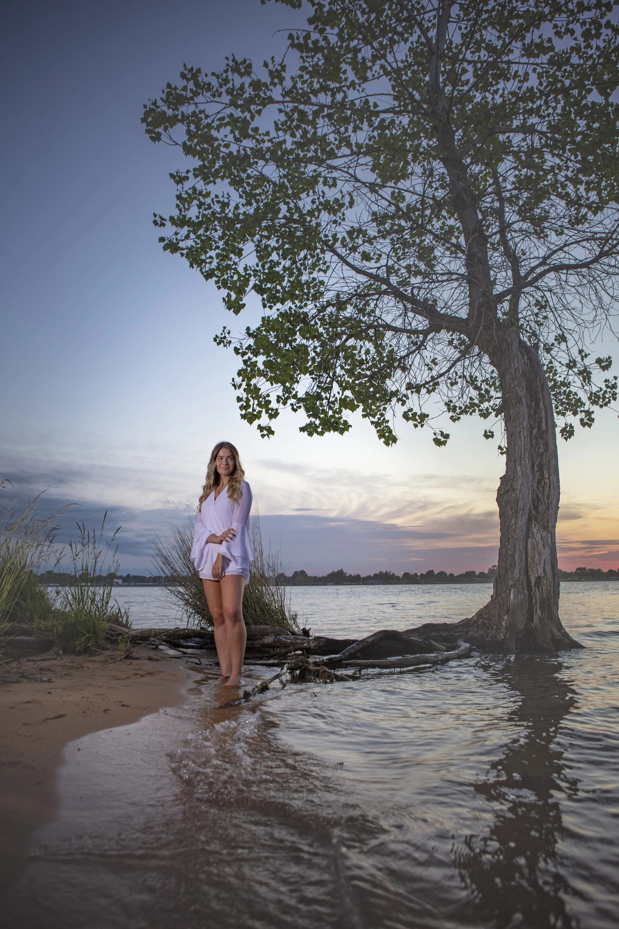 A woman in a white dress stands barefoot at the edge of a lake during sunset, next to a large tree with green leaves, on a sandy shoreline with some grass.
