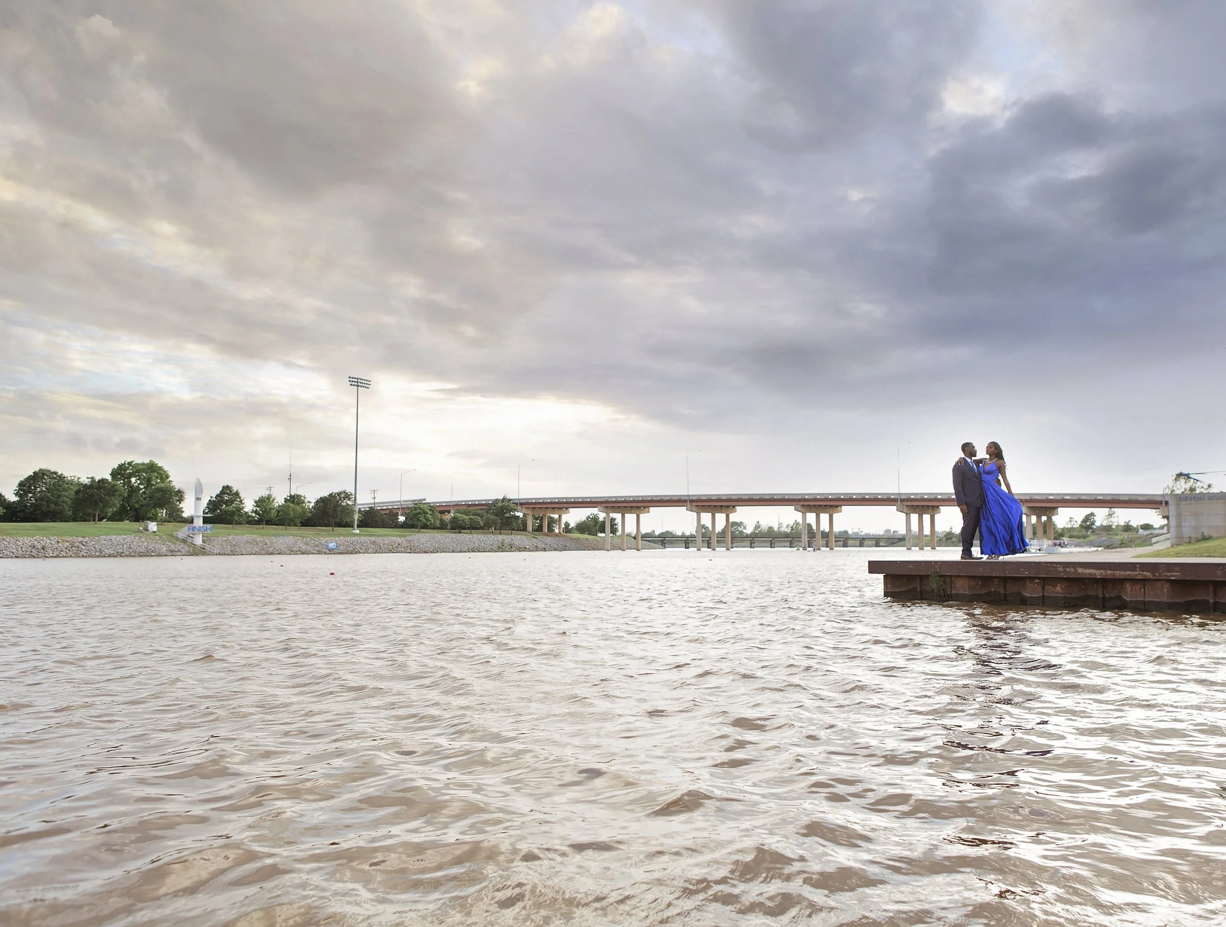 A couple dressed in formal attire standing on a dock by the river, with a bridge and cloudy sky in the background.