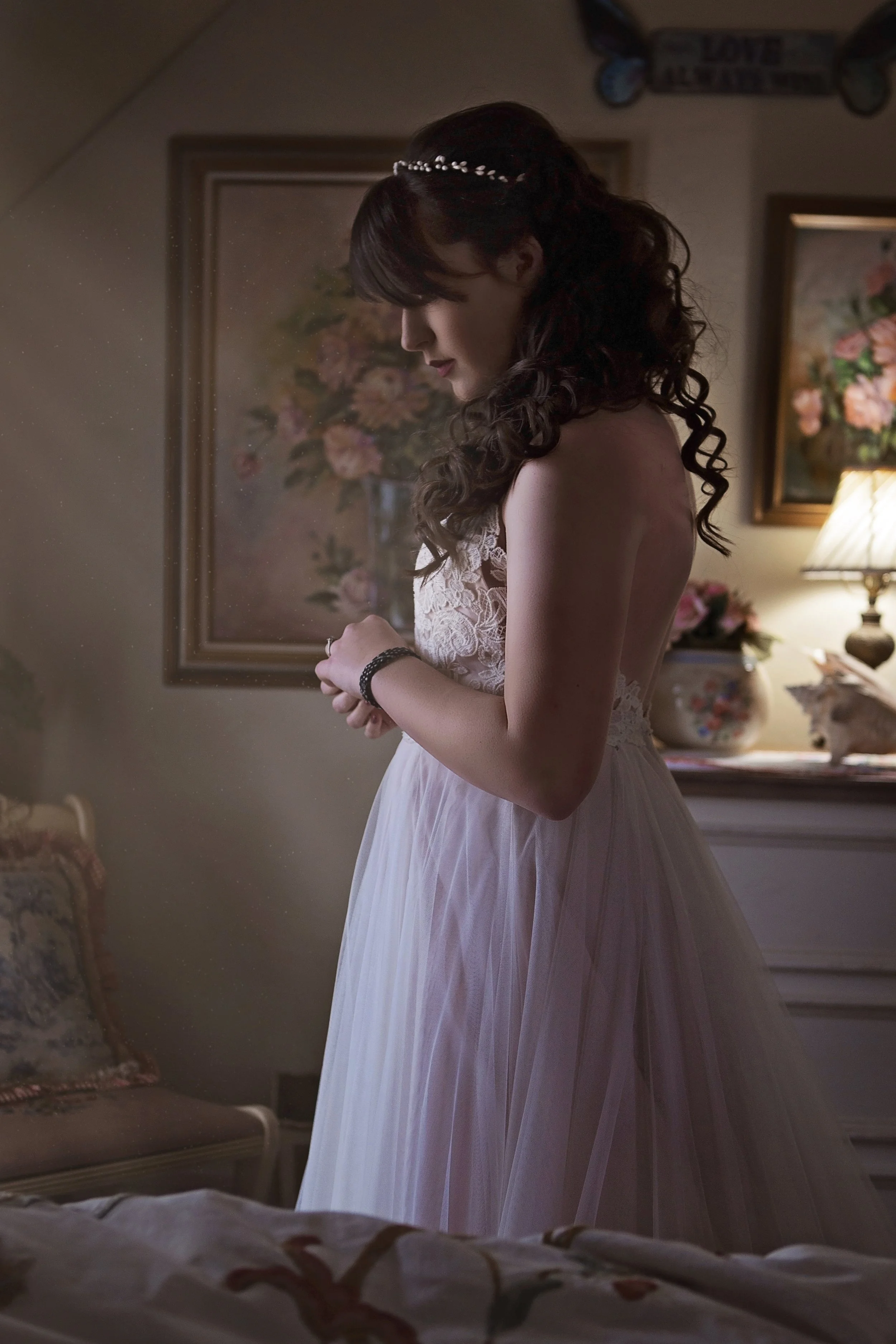 A woman in a white lace wedding dress with a tulle skirt, wearing a pearl headband, standing in a softly lit room with floral paintings and a lamp, clasping her hands.
