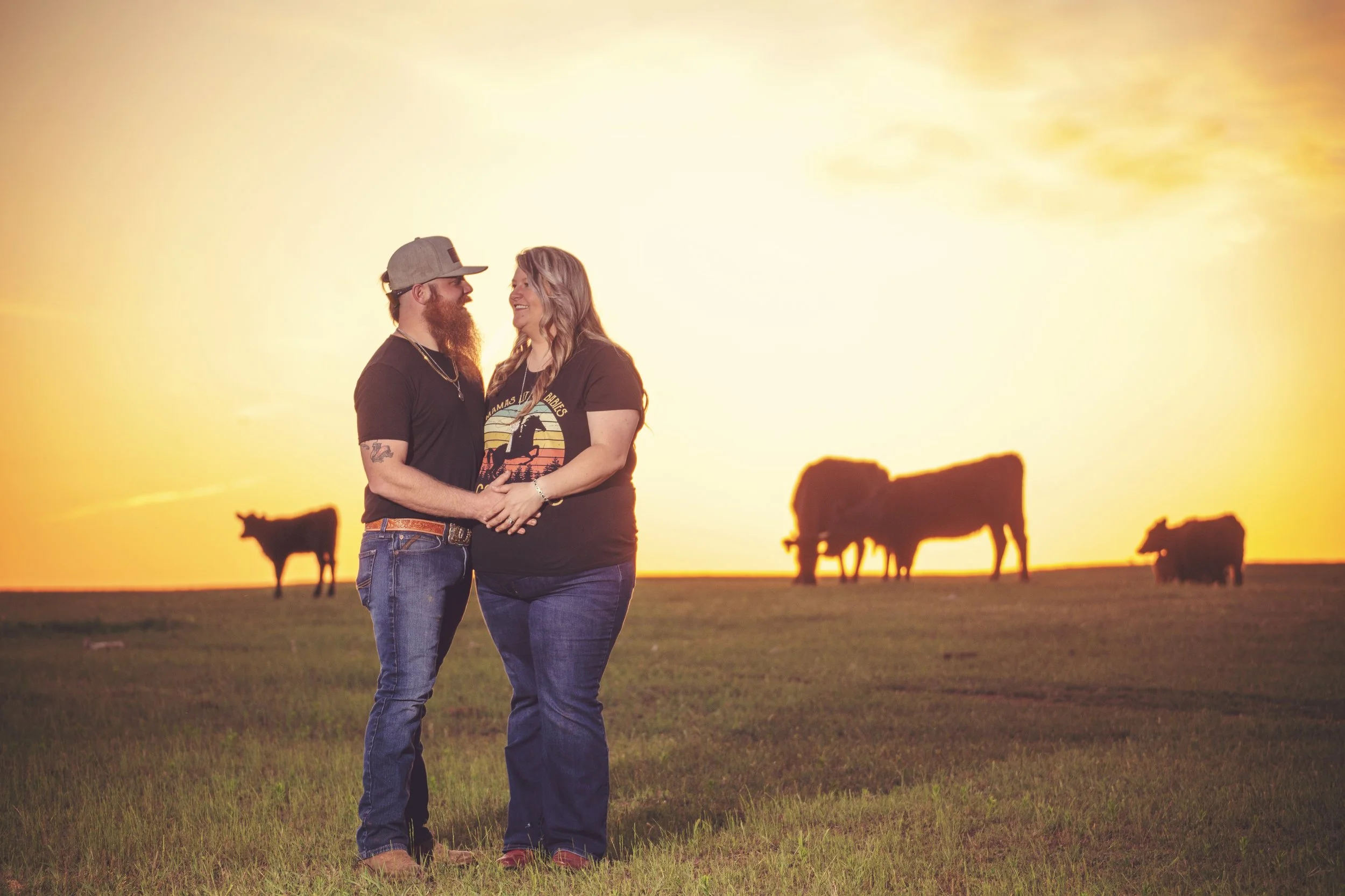 A couple standing close together holding hands in a grassy field at sunset, with a group of bison grazing in the background.