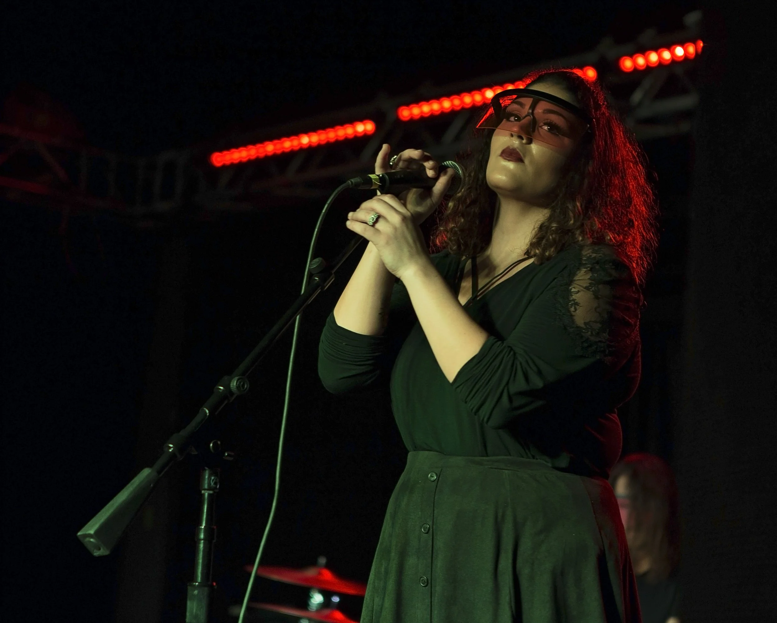 A woman with curly hair wearing glasses, a black top with lace sleeves, and a green skirt, singing into a microphone on stage with red lights in the background.