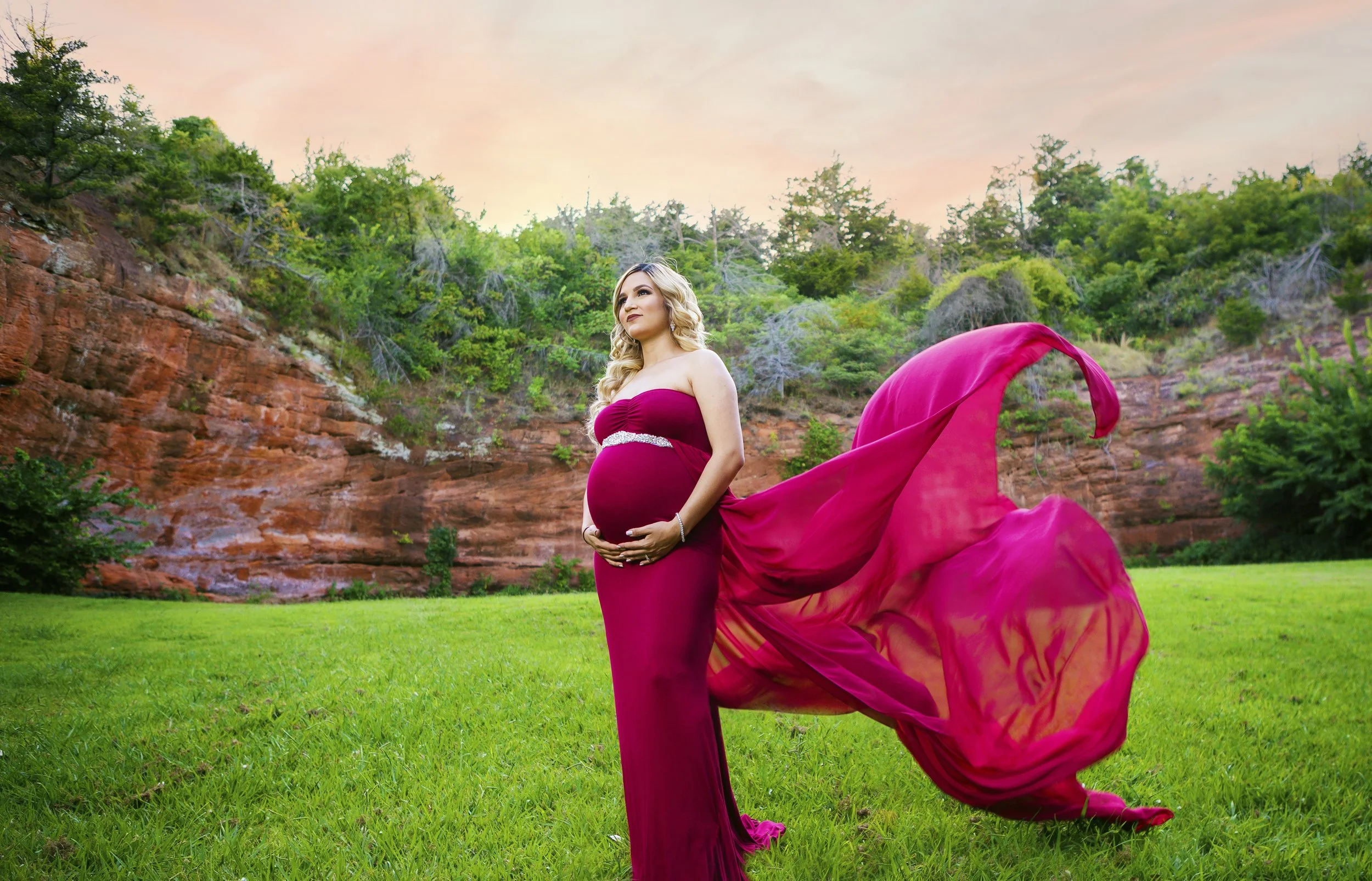 Pregnant woman in a red dress with a flowing train standing on green grass with trees and red rock cliffs in the background during sunset.