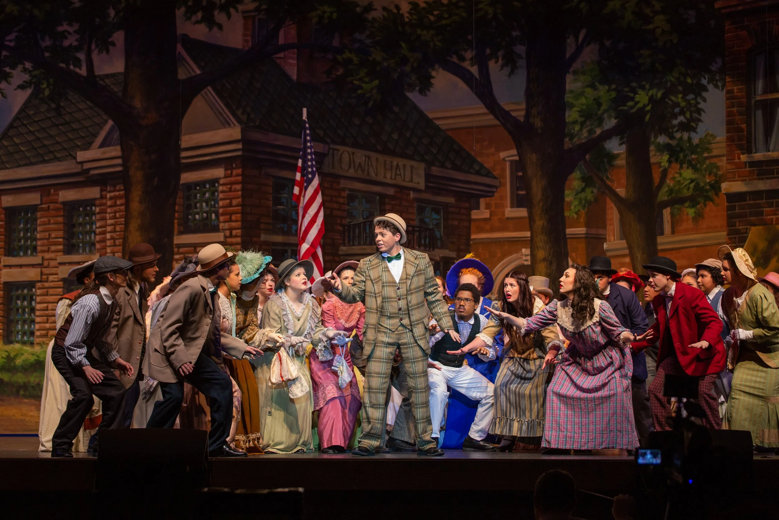 A theatrical performance on stage depicting a scene in a small town. A large group of actors dressed in vintage clothing, including hats, dresses, and suits, are gathered in front of a building labeled 'Town Hall'. The scene appears lively, with some