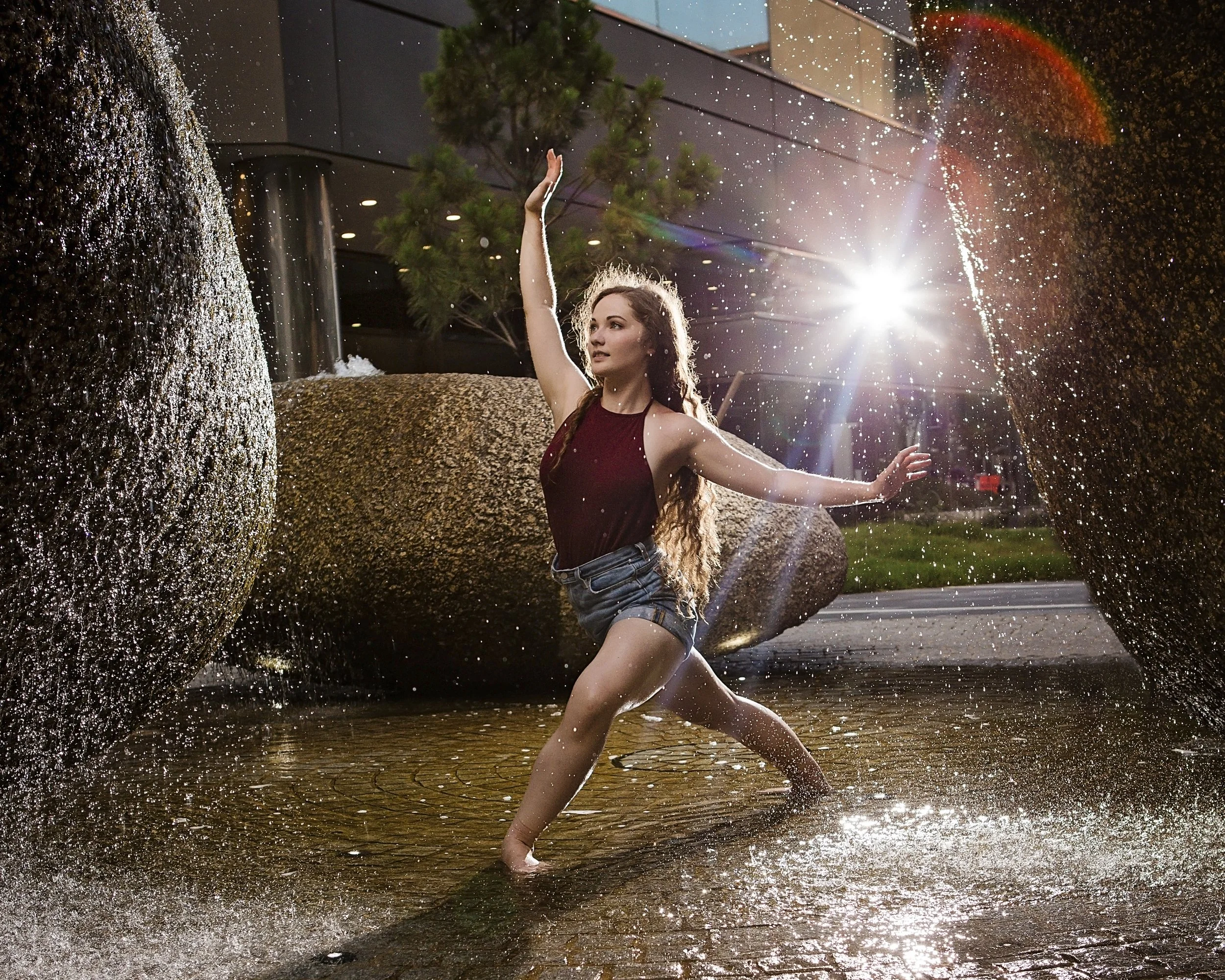 A young woman with long, curly hair wading through a small water fountain at sunset, with the sun shining brightly in the background and a rainbow forming in the sky.