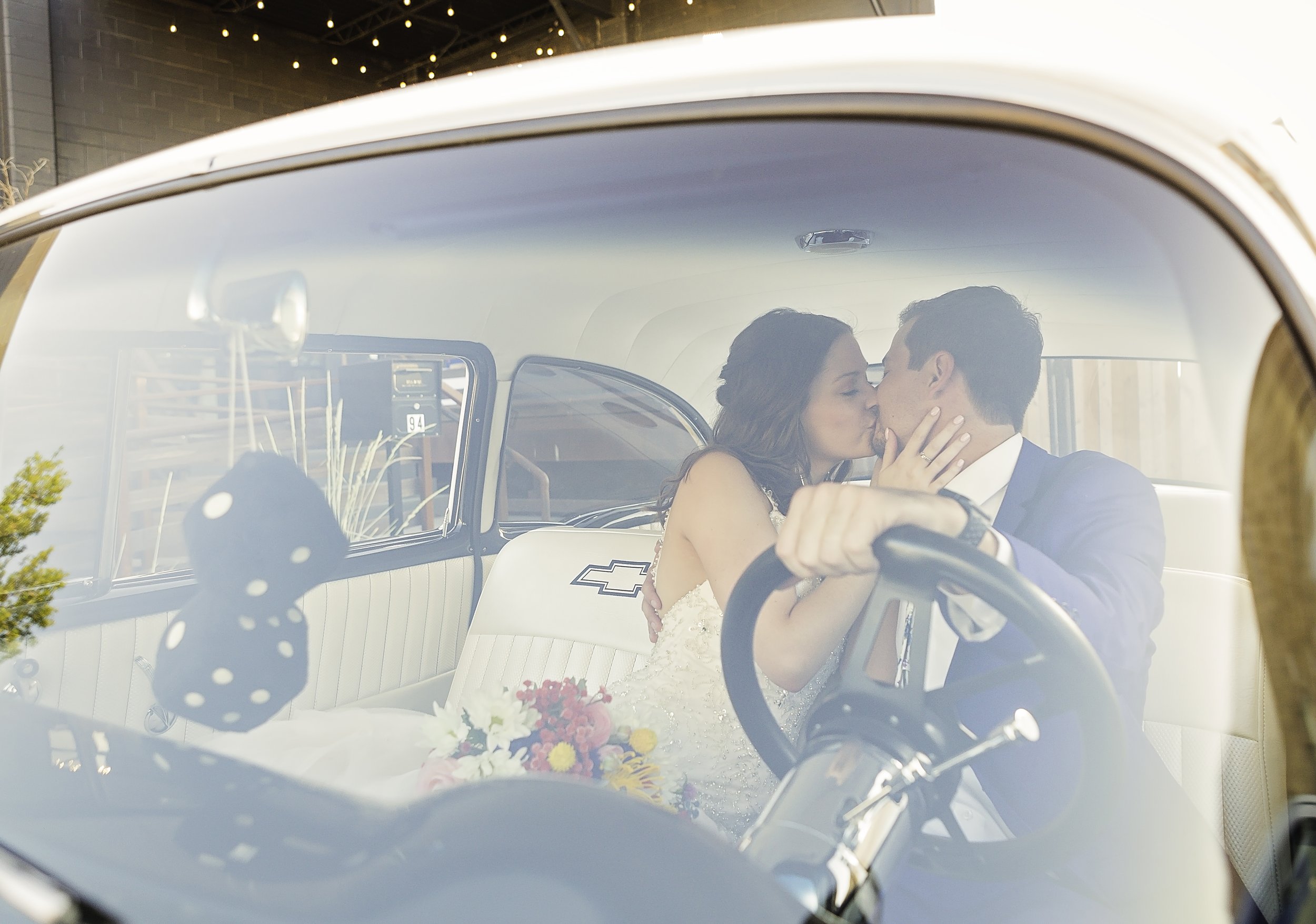 A bride and groom sharing a kiss inside a vintage car, with the bride holding a bouquet of flowers and a Black polka-dotted die hanging from the rearview mirror.