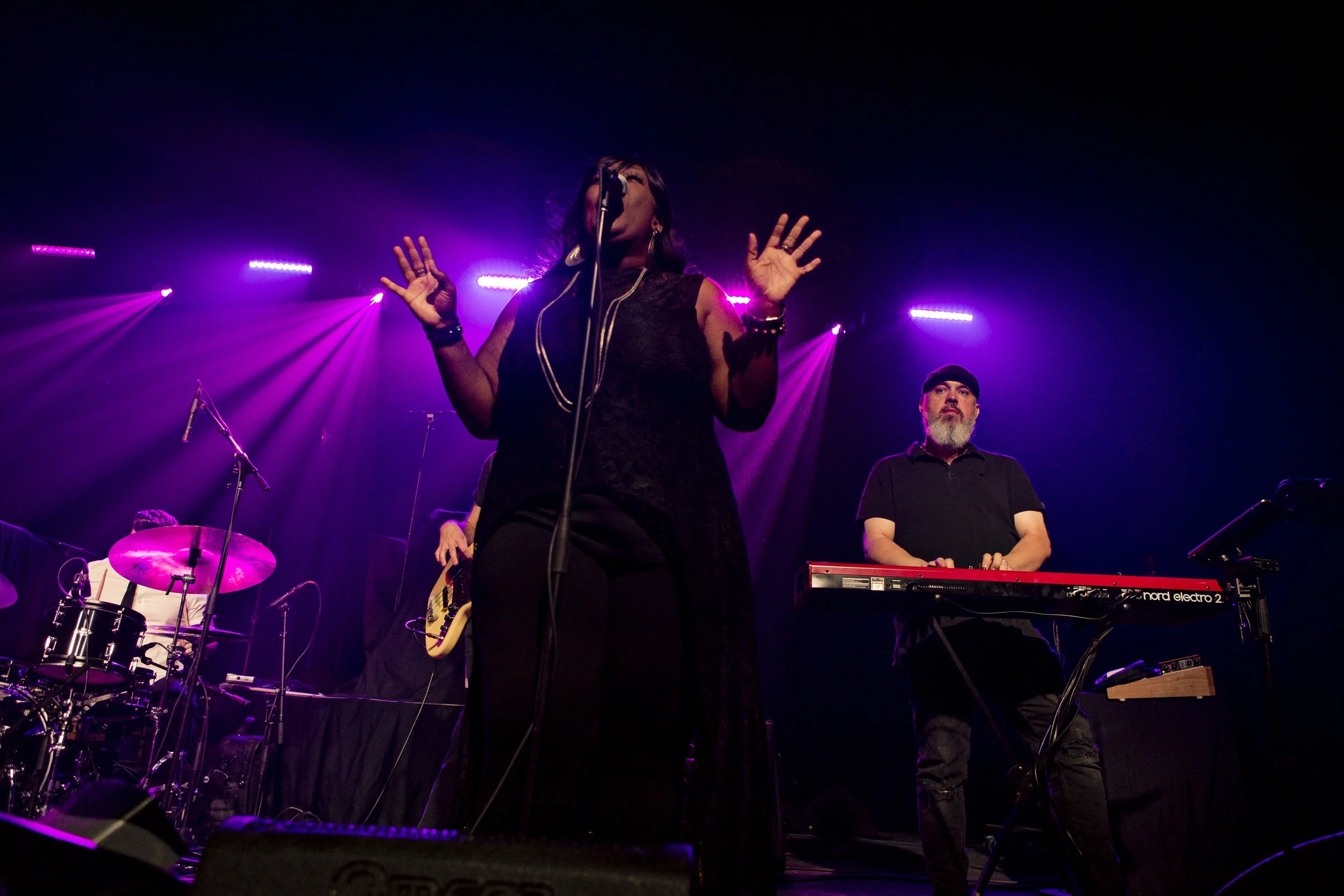 Musicians performing on stage under purple and pink lighting, including a female singer with hands raised, a keyboard player, a guitarist, and a drummer partially visible on the left.