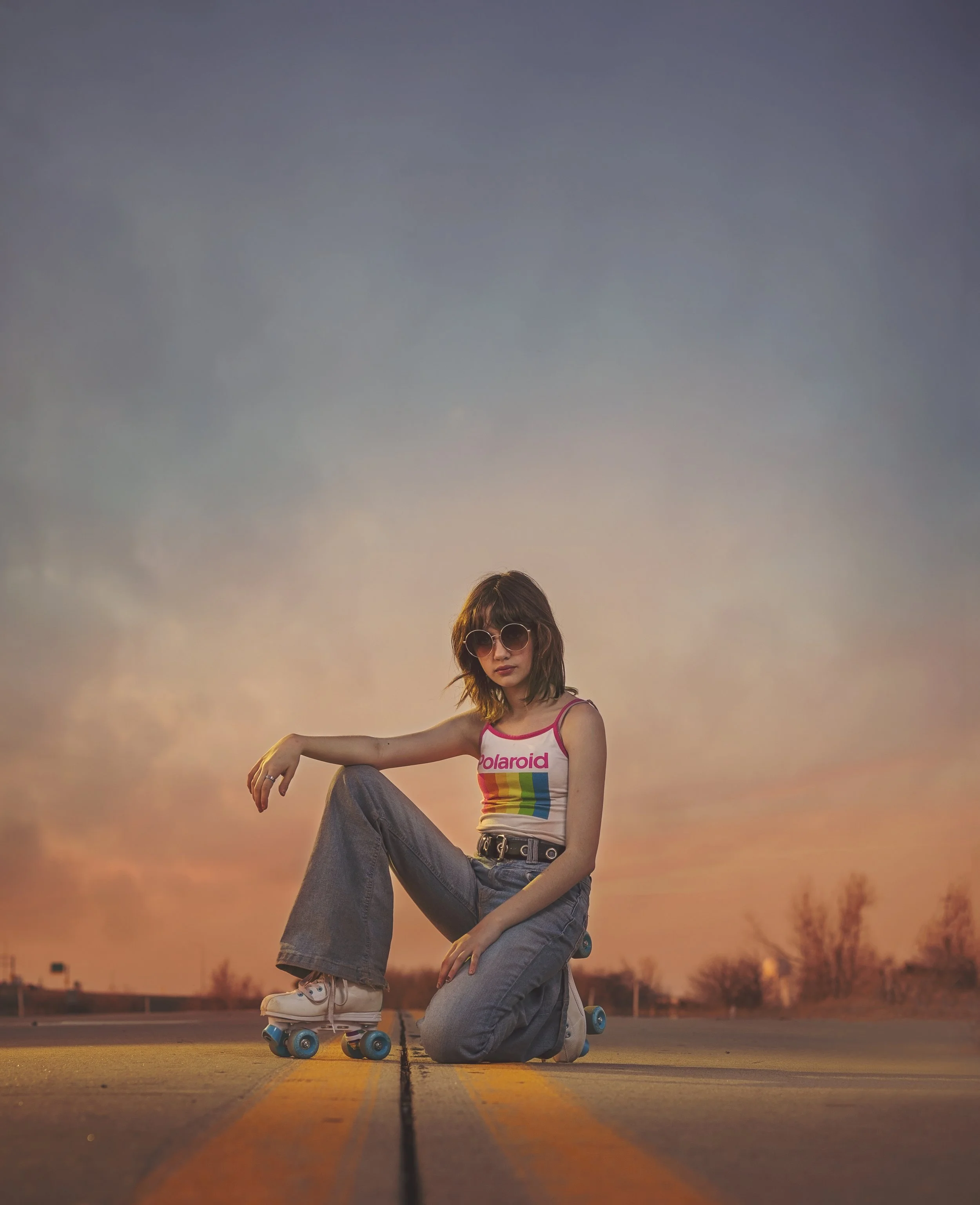 A young woman wearing sunglasses, a Polaroid tank top, and jeans, posing with roller skates on a road at sunset.