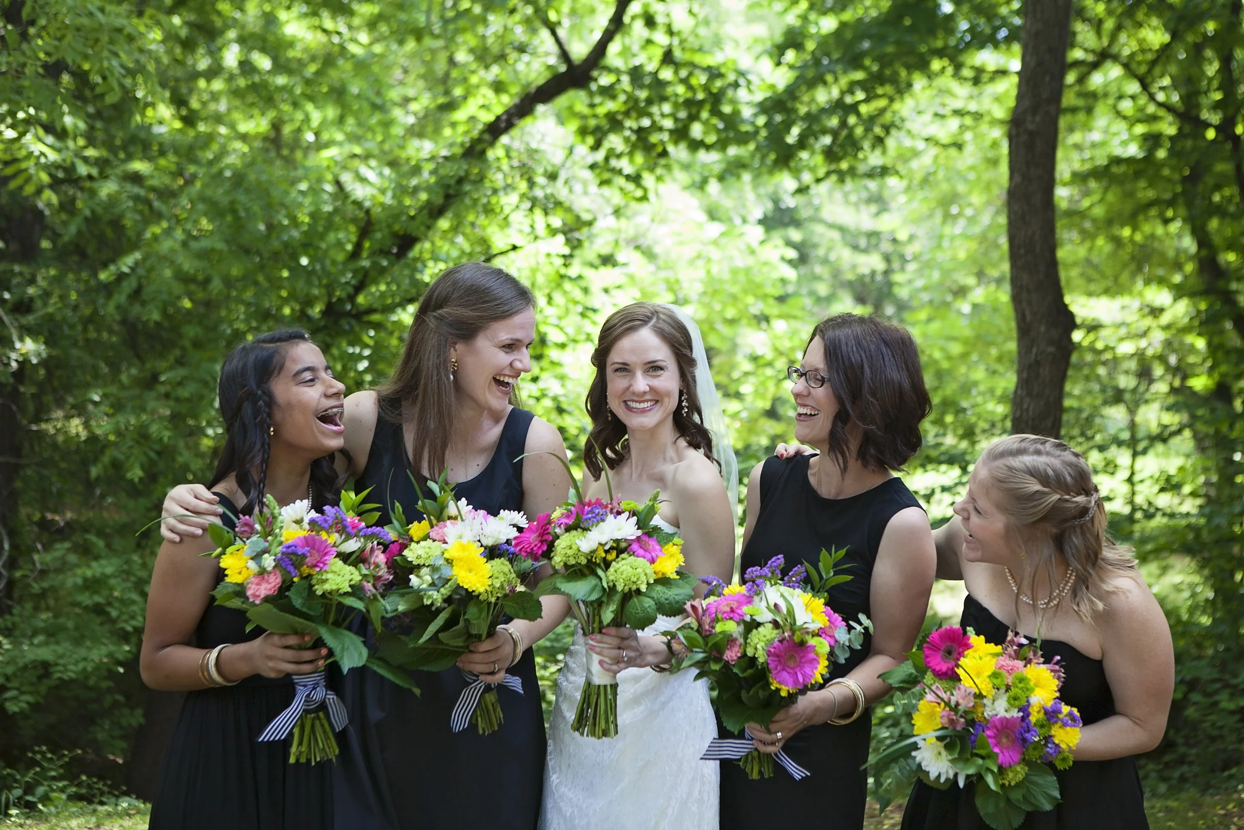 Group of women, including a bride in a white dress, holding bouquets of colorful flowers, standing outdoors in a green wooded area, smiling and laughing together.