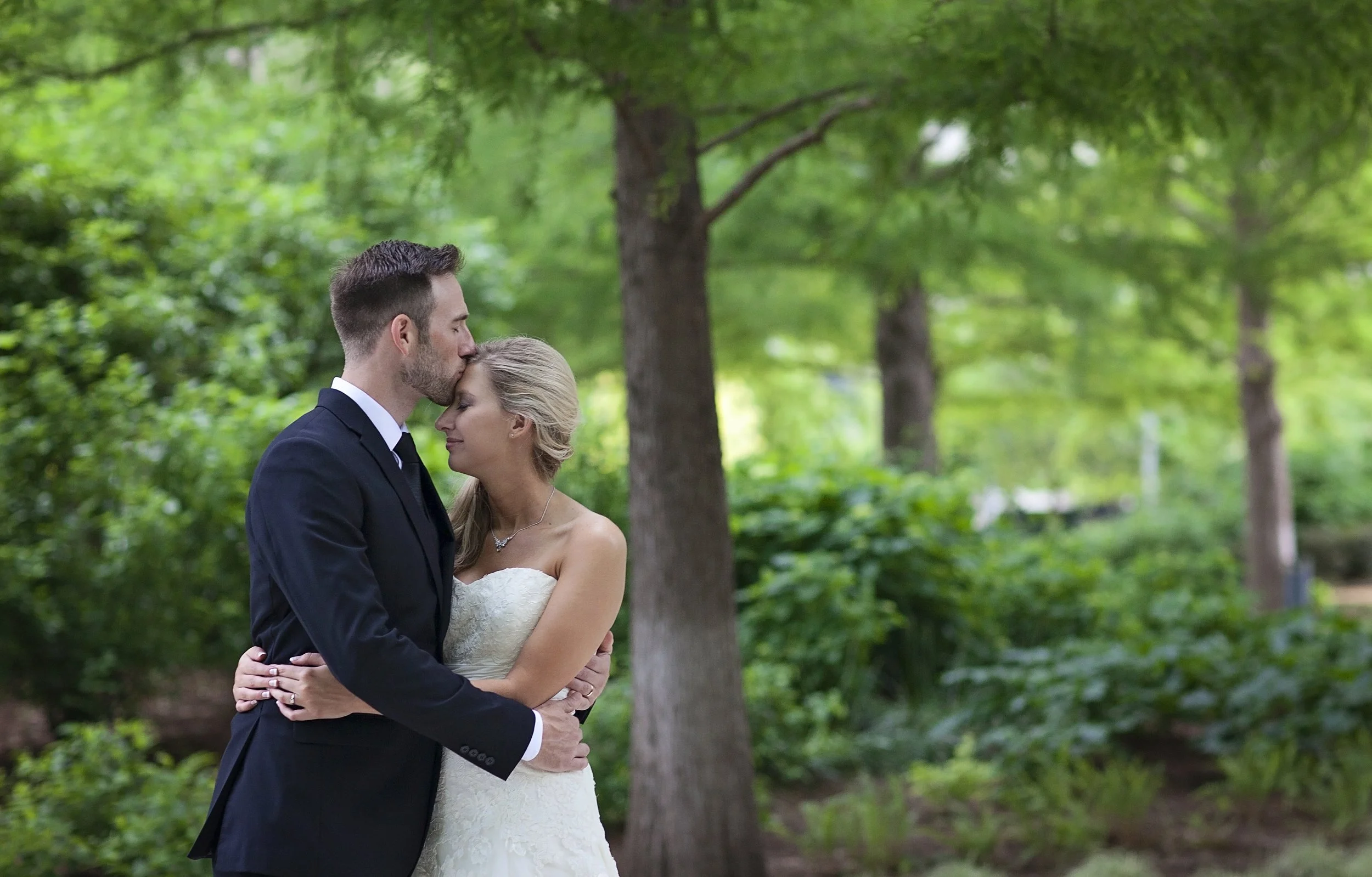 A bride and groom embrace in a lush green outdoor setting, with the groom kissing the bride's forehead.