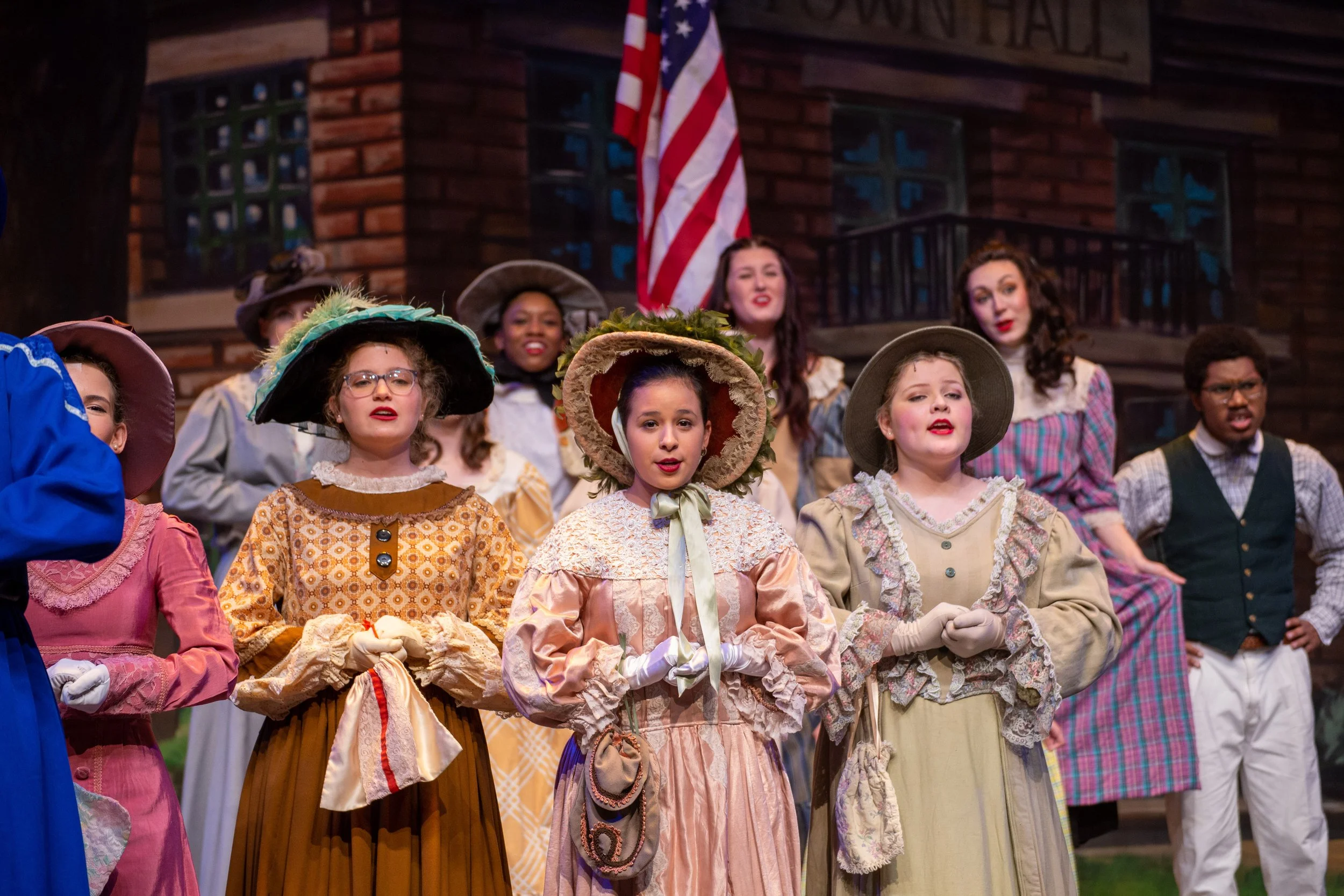 Young girls dressed in period costumes singing on stage during a theatrical performance, with a wooden building and American flag in background.