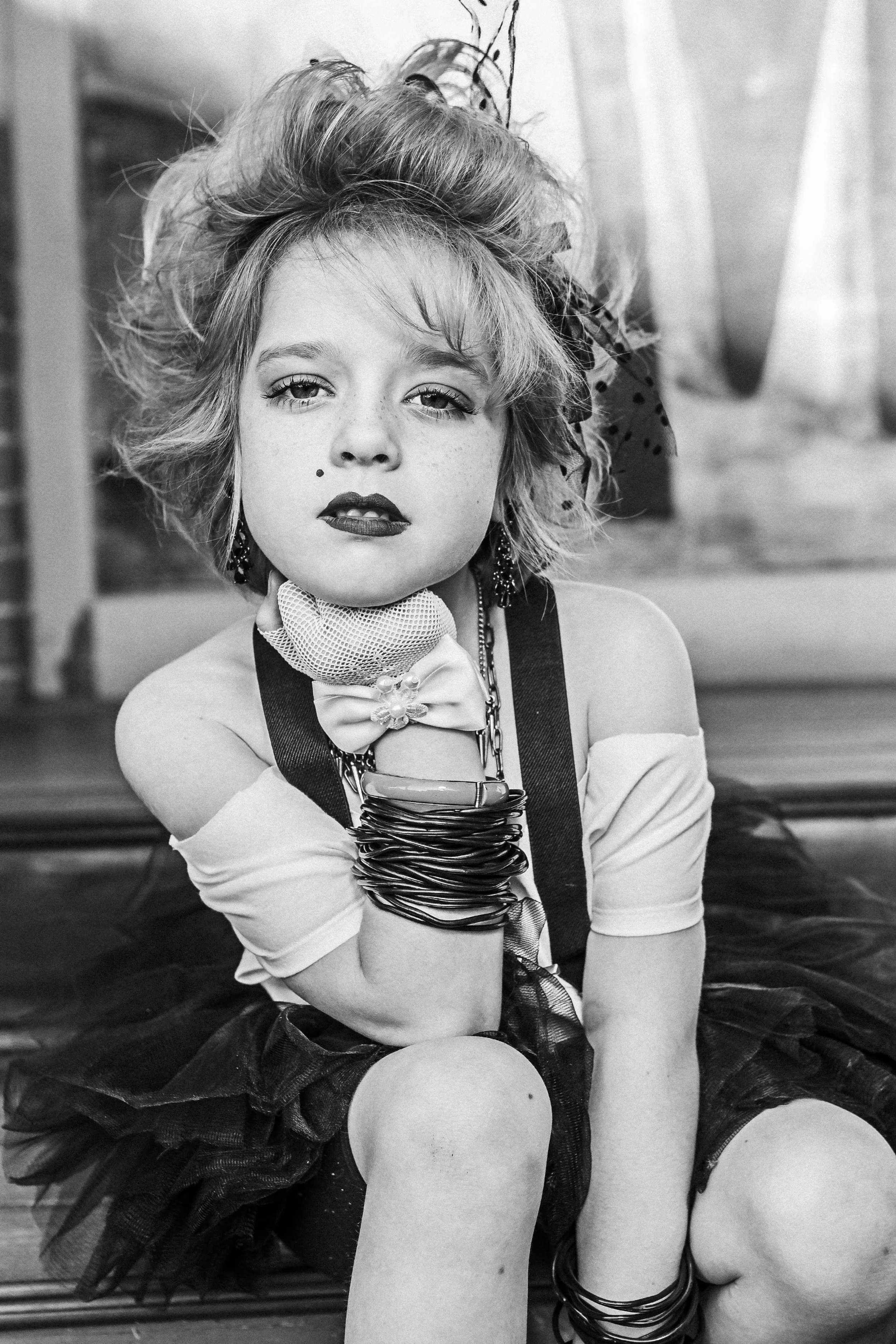 Black and white photo of a young woman with wild, tousled hair, wearing makeup, earrings, and multiple bracelets, resting her chin on her hand with a serious expression.