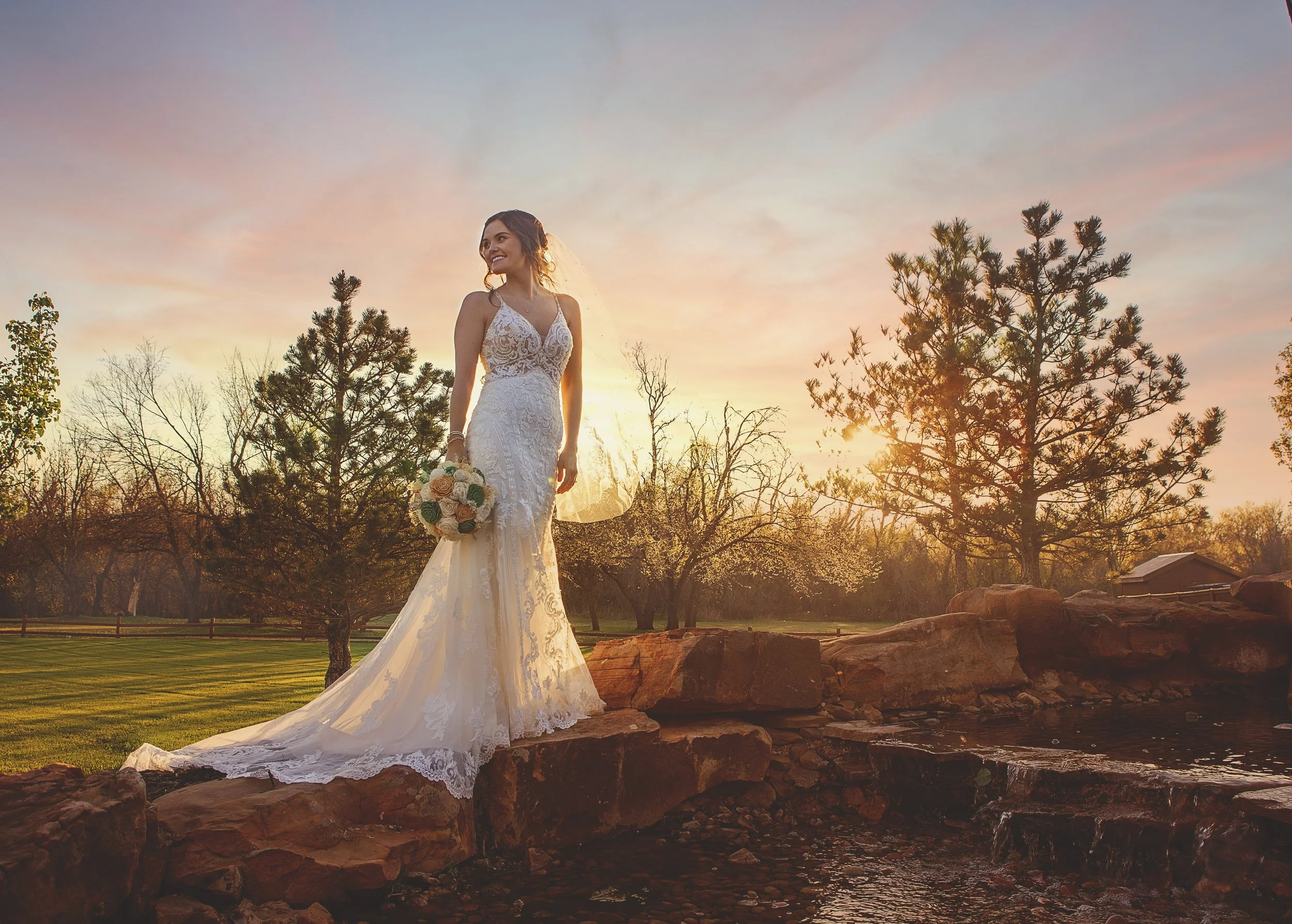 A bride in a white lace wedding dress holding a bouquet of roses standing on rocks near a small waterfall during sunset, with trees and open field in the background.
