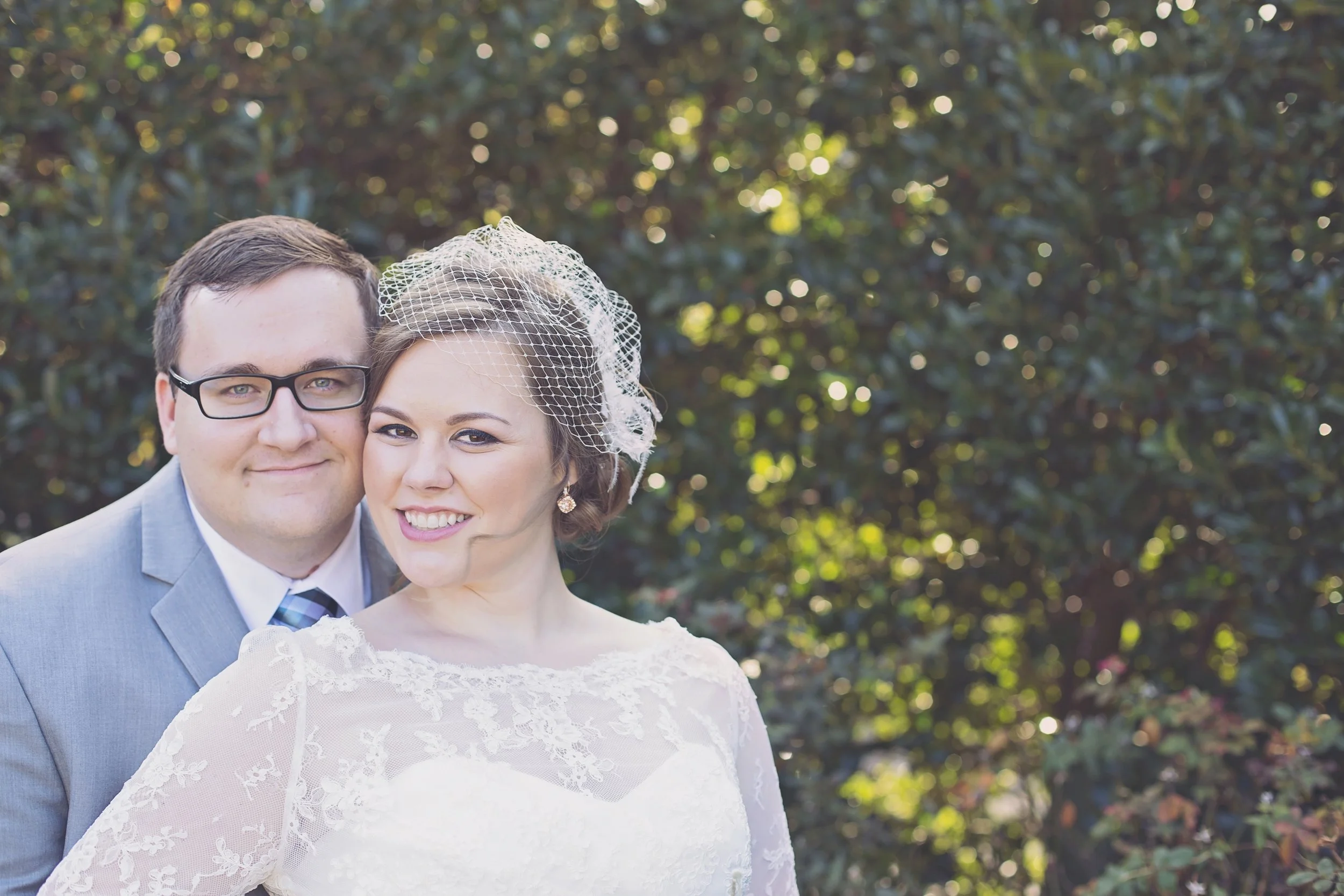 Couple on their wedding day outdoors, the groom wearing a light gray suit and the bride in a white lace wedding dress and birdcage veil, standing in front of a leafy background.