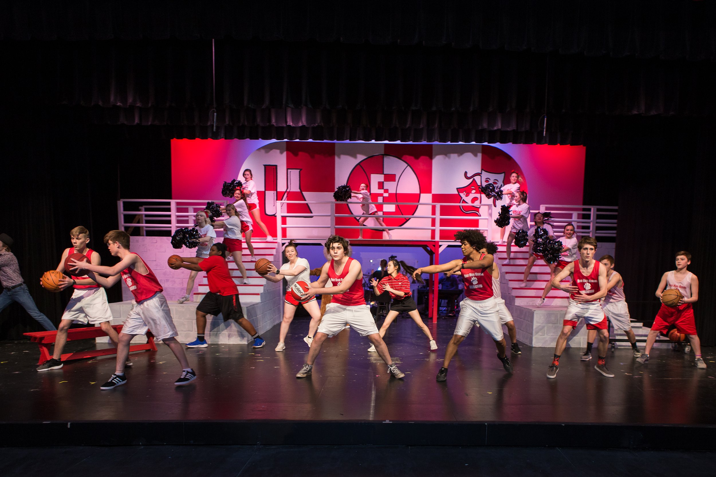 Children performing a basketball-themed musical on stage, with dancers in cheerleading outfits and a colorful school emblem background.
