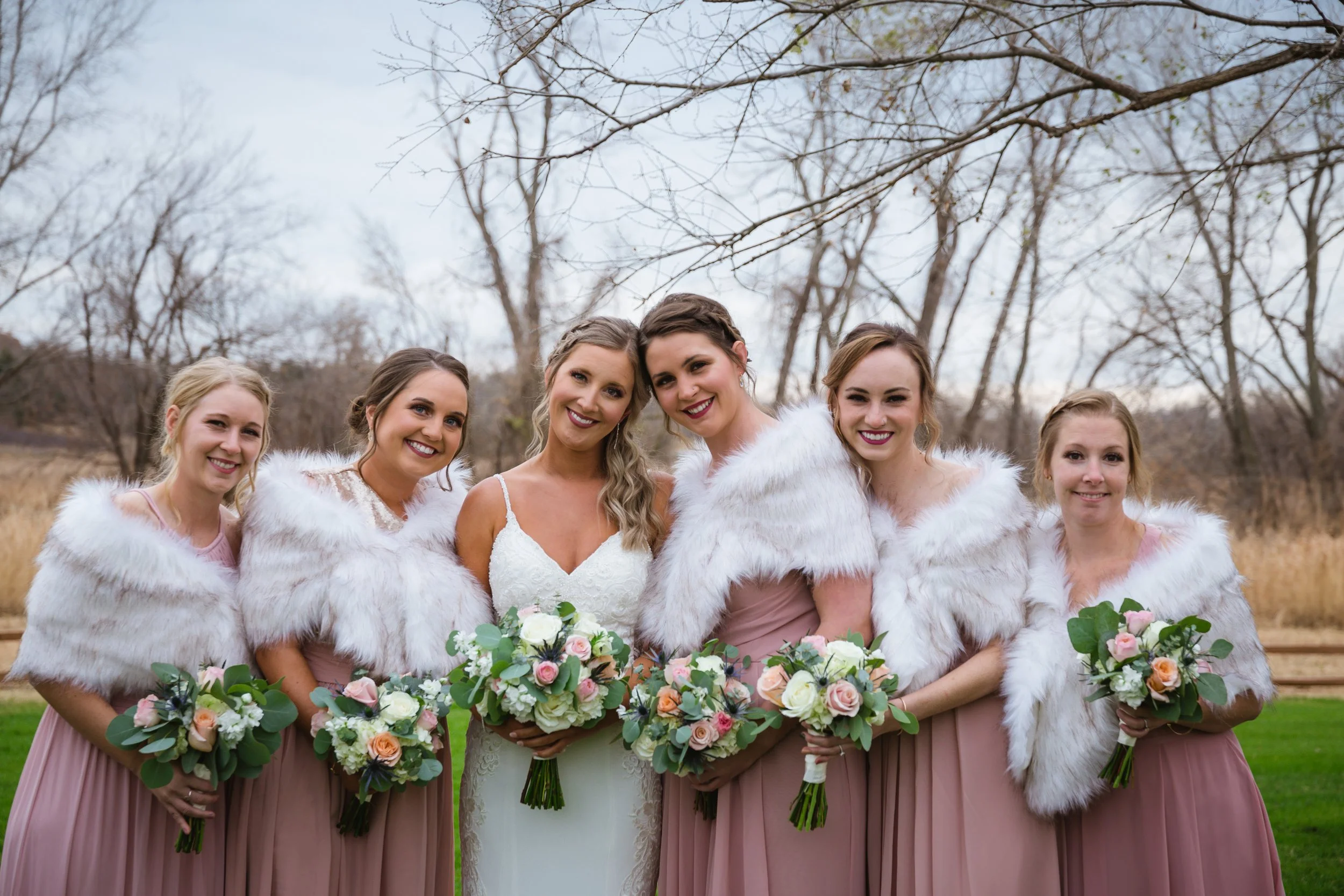 Bride and five bridesmaids in outdoor setting, dressed in pink dresses with white fur shawls, holding bouquets of pink, white, and green flowers.