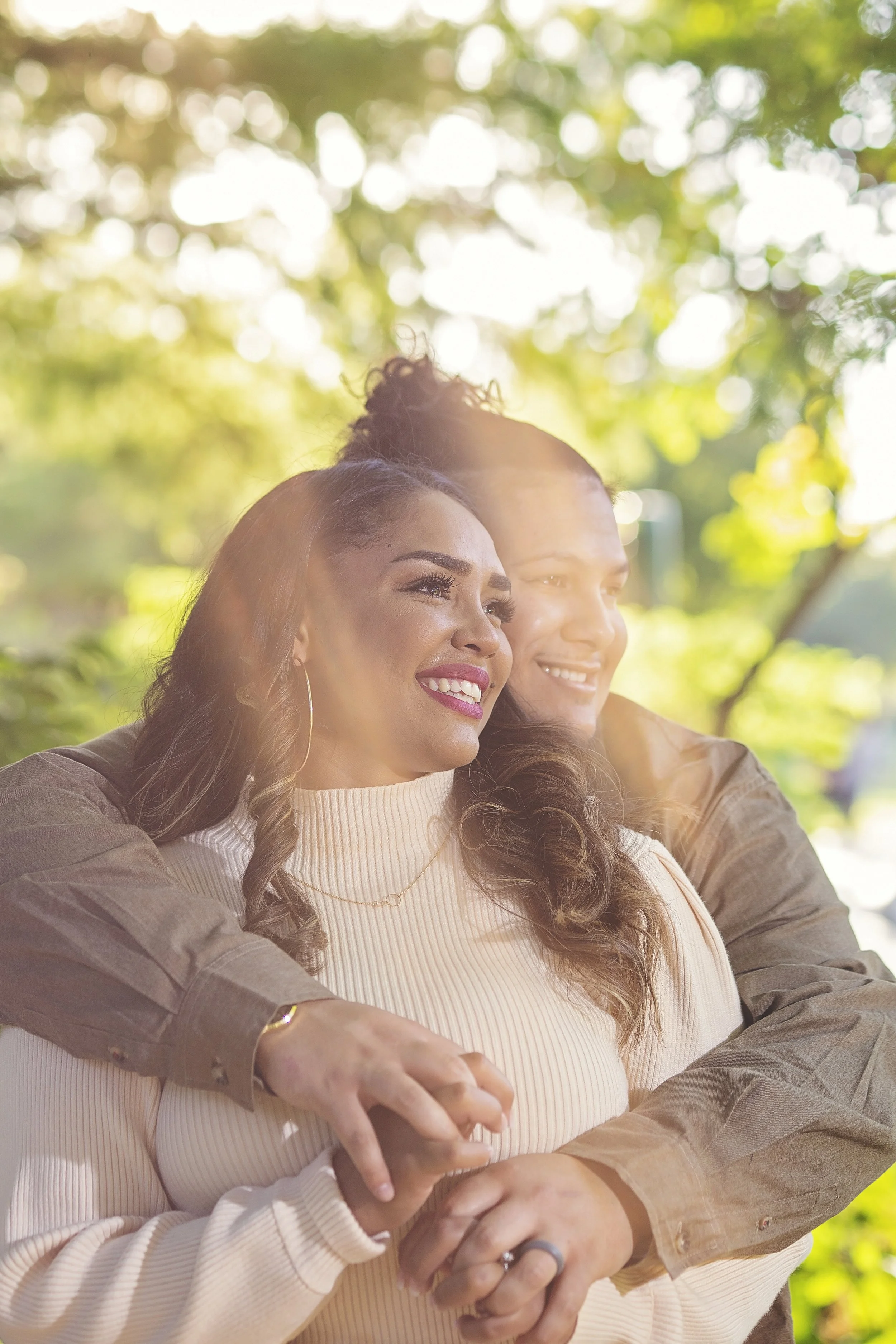 A couple hugging outdoors on a sunny day with green trees in the background.