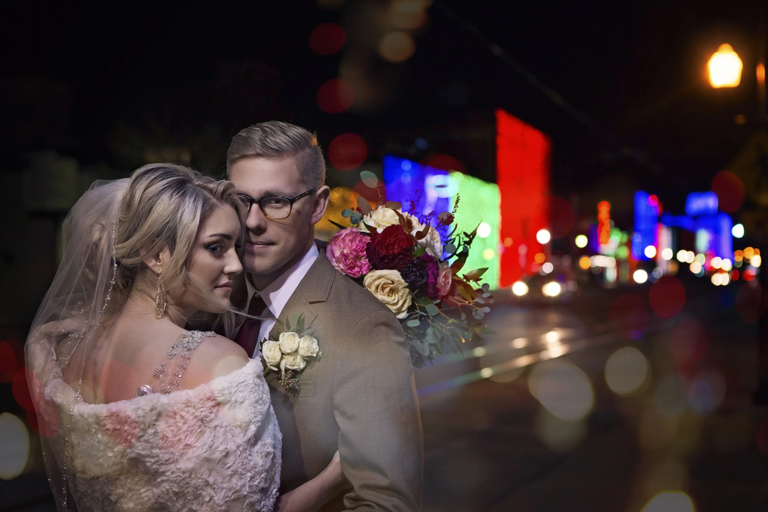 A newlywed couple at night in front of colorful city lights, with the groom wearing glasses and a suit, and the bride in a wedding dress with a veil, holding a bouquet of flowers.