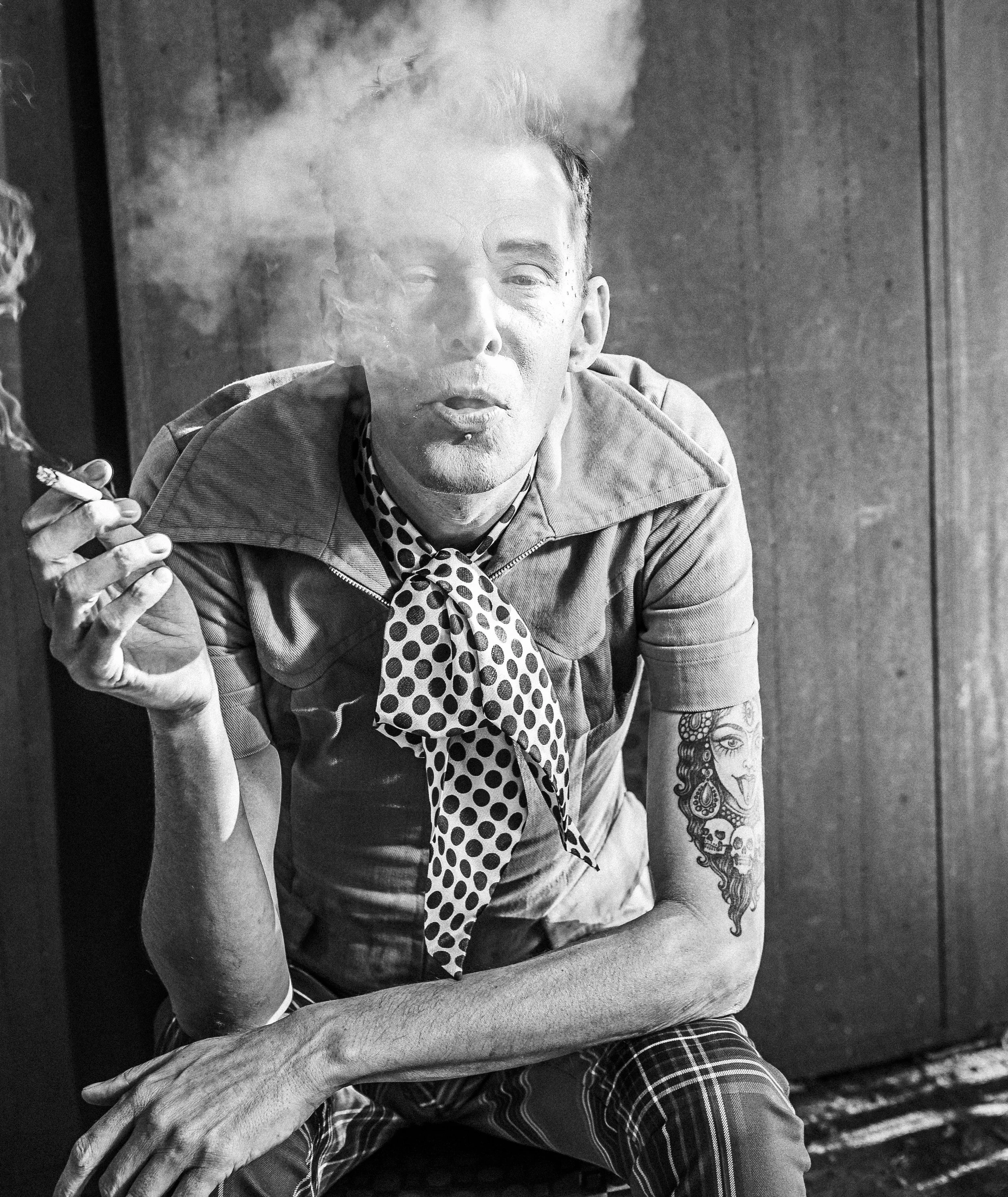 A man with tattoos on his arm smoking a cigarette, wearing a polka dot scarf and jacket, sitting against a wooden wall, with smoke around his face.