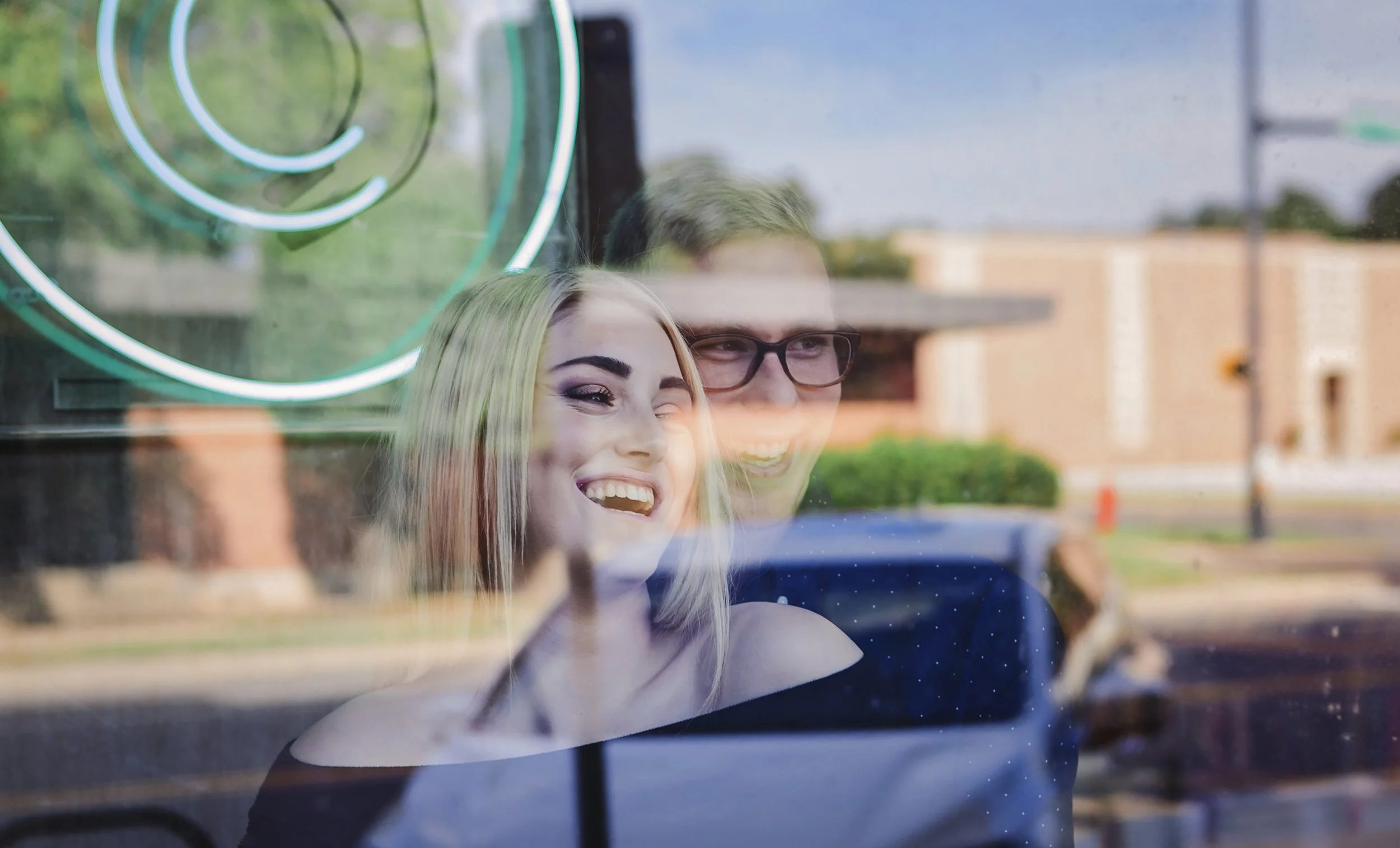 Engagement image looking out of a window with neon sign, outdoors during daytime