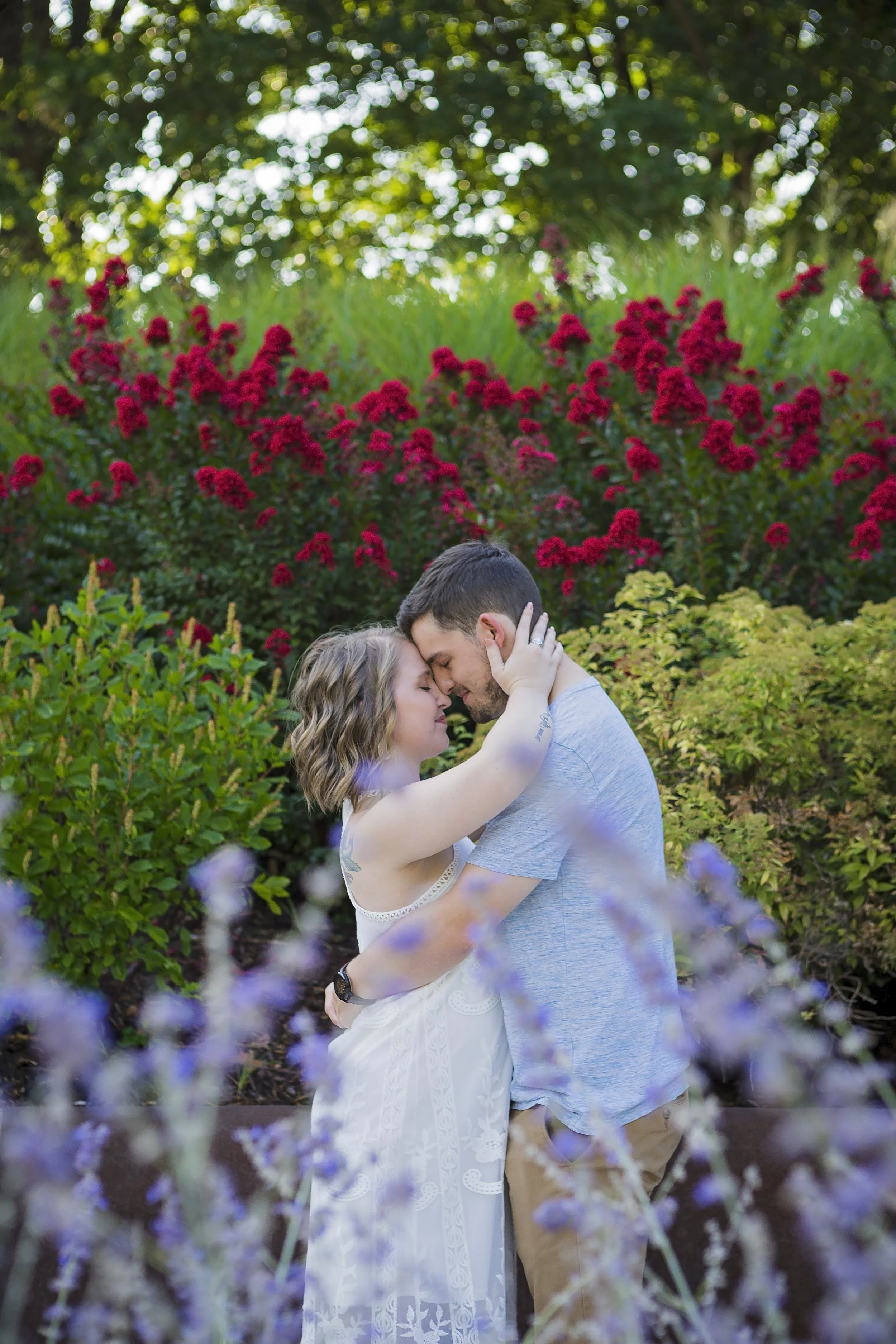 A couple embraces tenderly in a garden with vibrant pink and purple flowers and green foliage, with sunlight filtering through trees in the background.