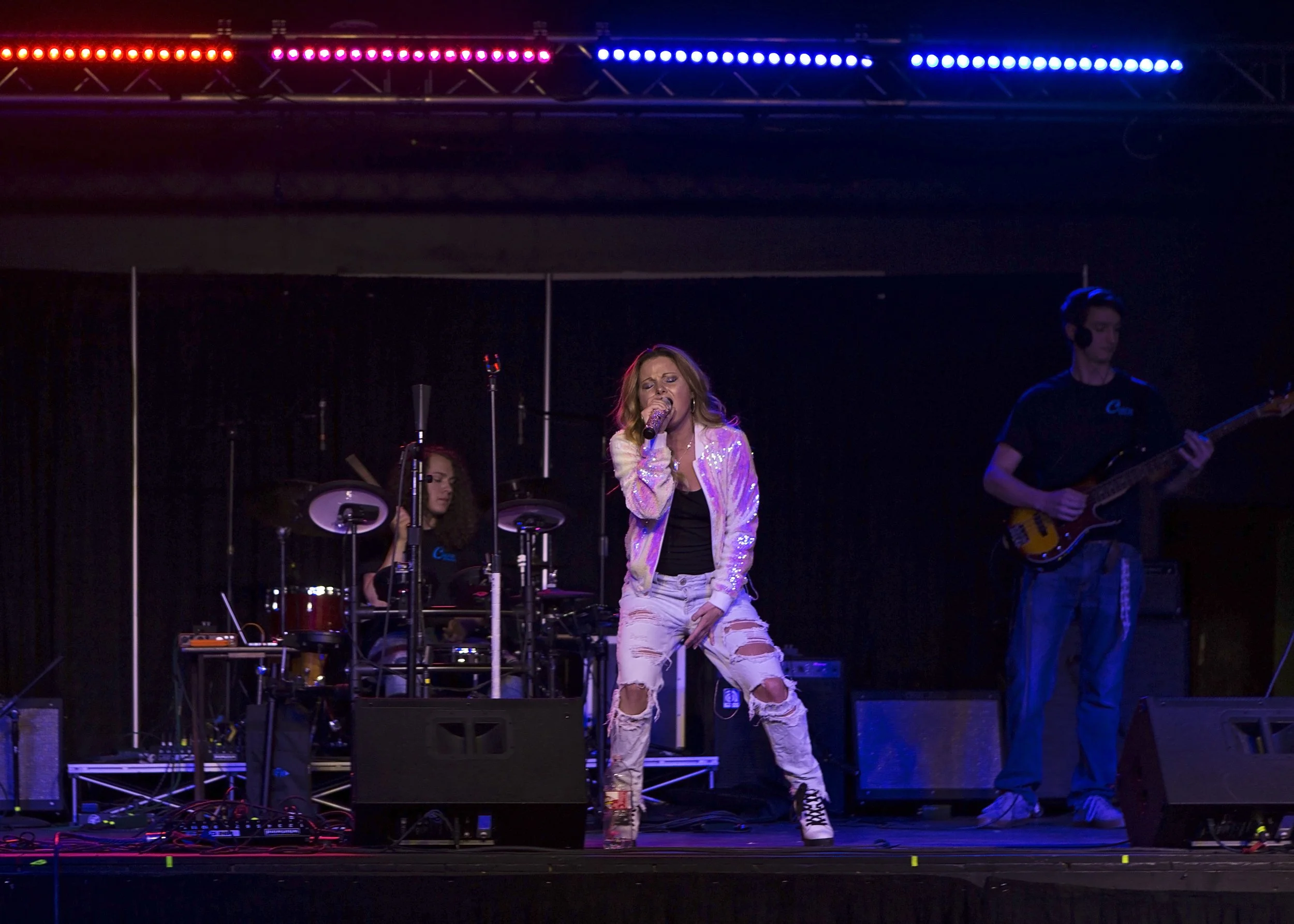 Female singer with long red hair in a white, shiny jacket and ripped jeans performs on stage with two musicians, a drummer and a bassist, under colorful stage lights.