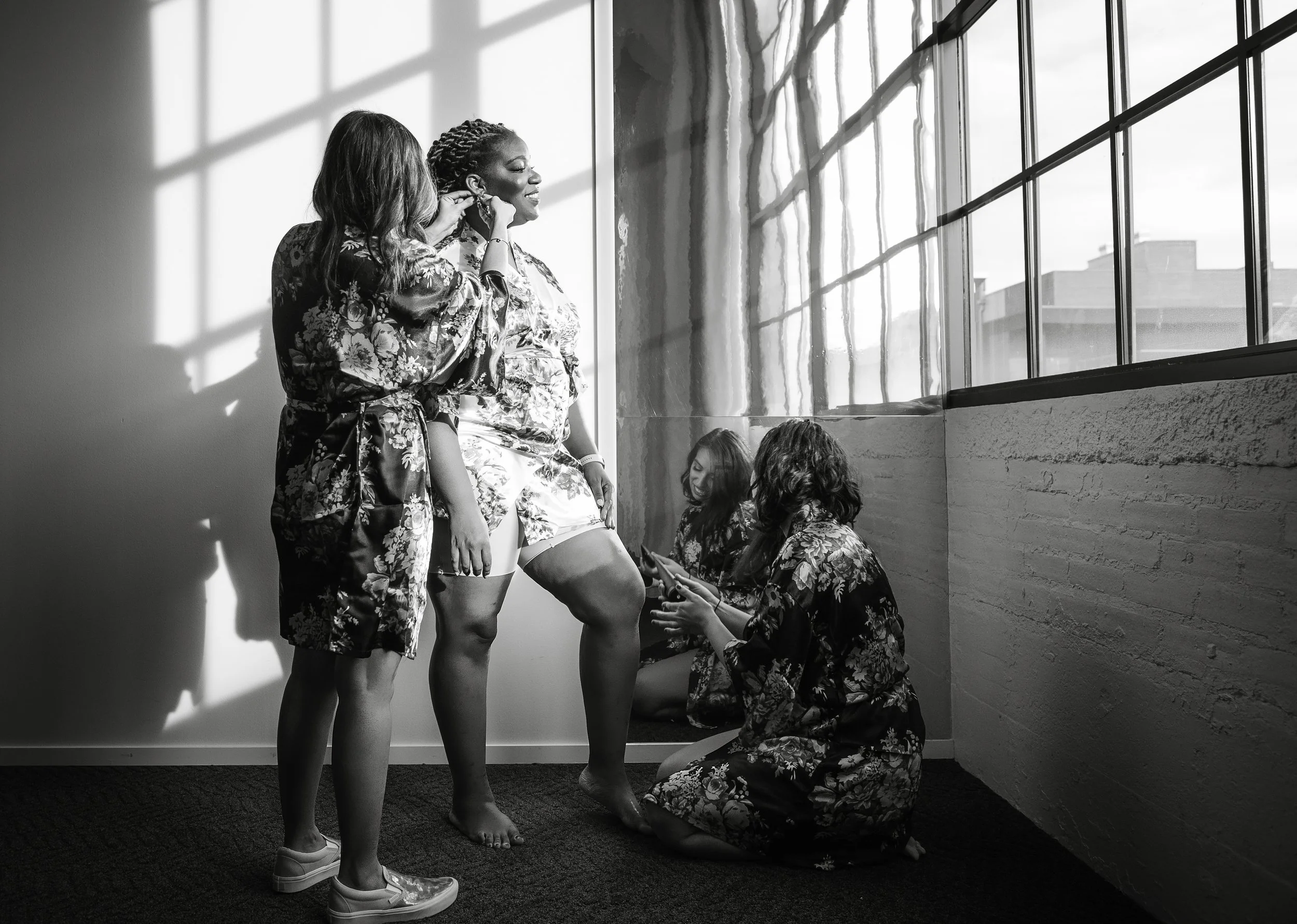 Bridal party getting ready in  floral robes preparing a bride in a bridal suite, with natural sunlight coming through large windows.