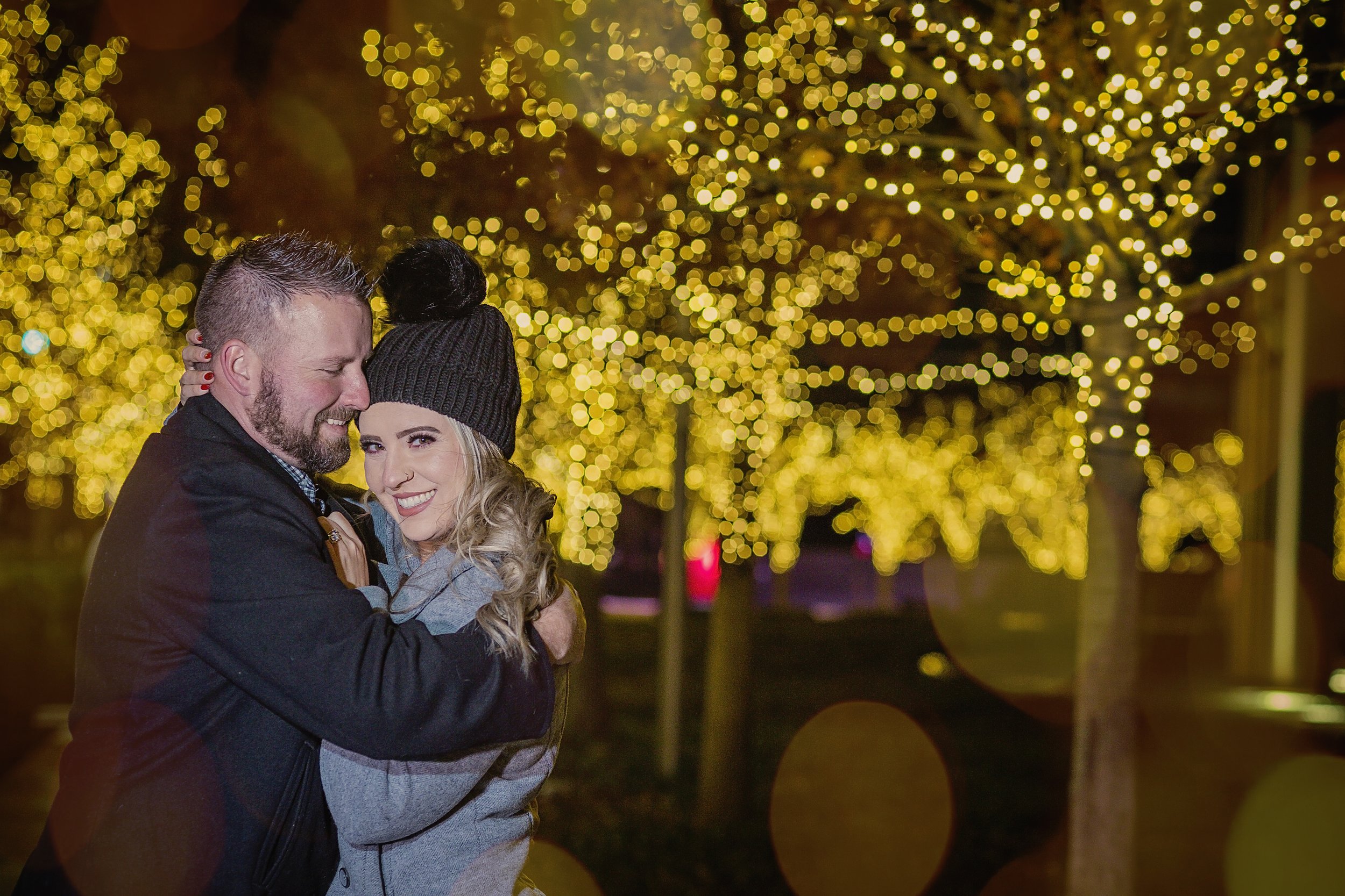 A couple hugging and smiling at night with trees decorated with yellow Christmas lights in the background.