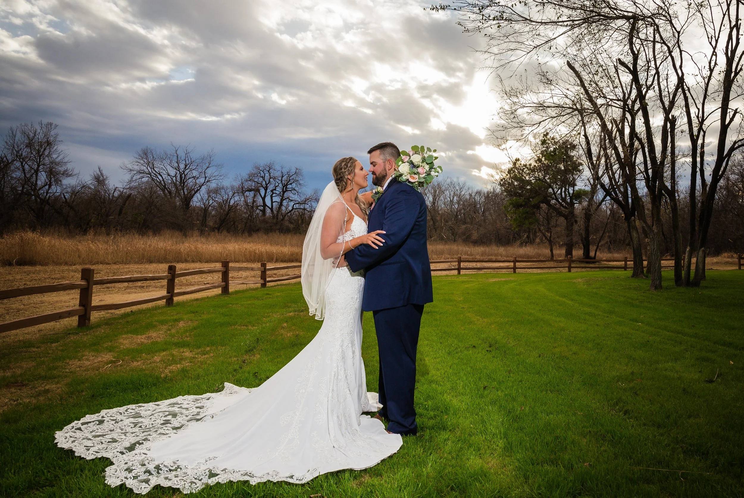 A bride and groom embrace on a grassy field outdoors with trees and a cloudy sky in the background. The bride wears a white lace wedding gown with a long train and veil, and the groom wears a navy suit holding a bouquet of flowers.