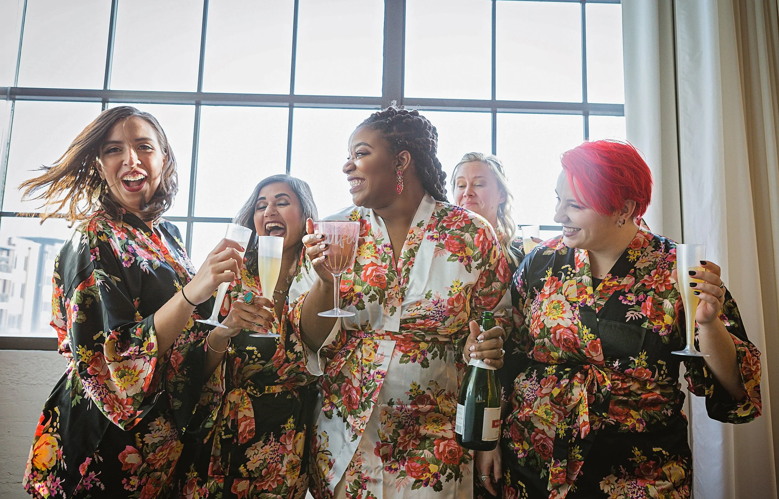 Group of women celebrating with drinks, wearing floral print robes and pajamas, indoors near large windows.