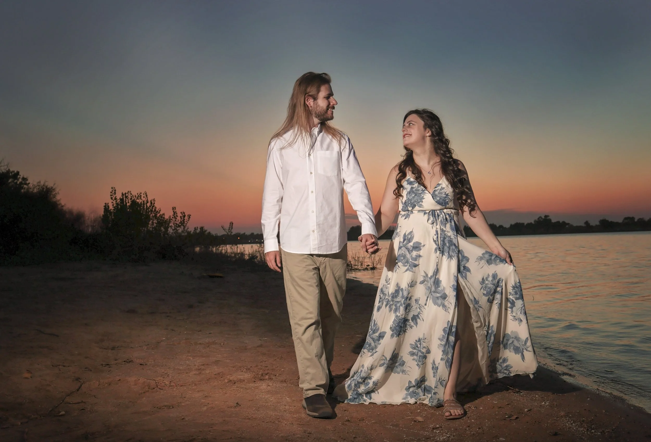 Lake beach engagement session of this couple holding hands and walking on a beach at sunset, with the woman wearing a long floral dress and the man in a white shirt and beige pants.