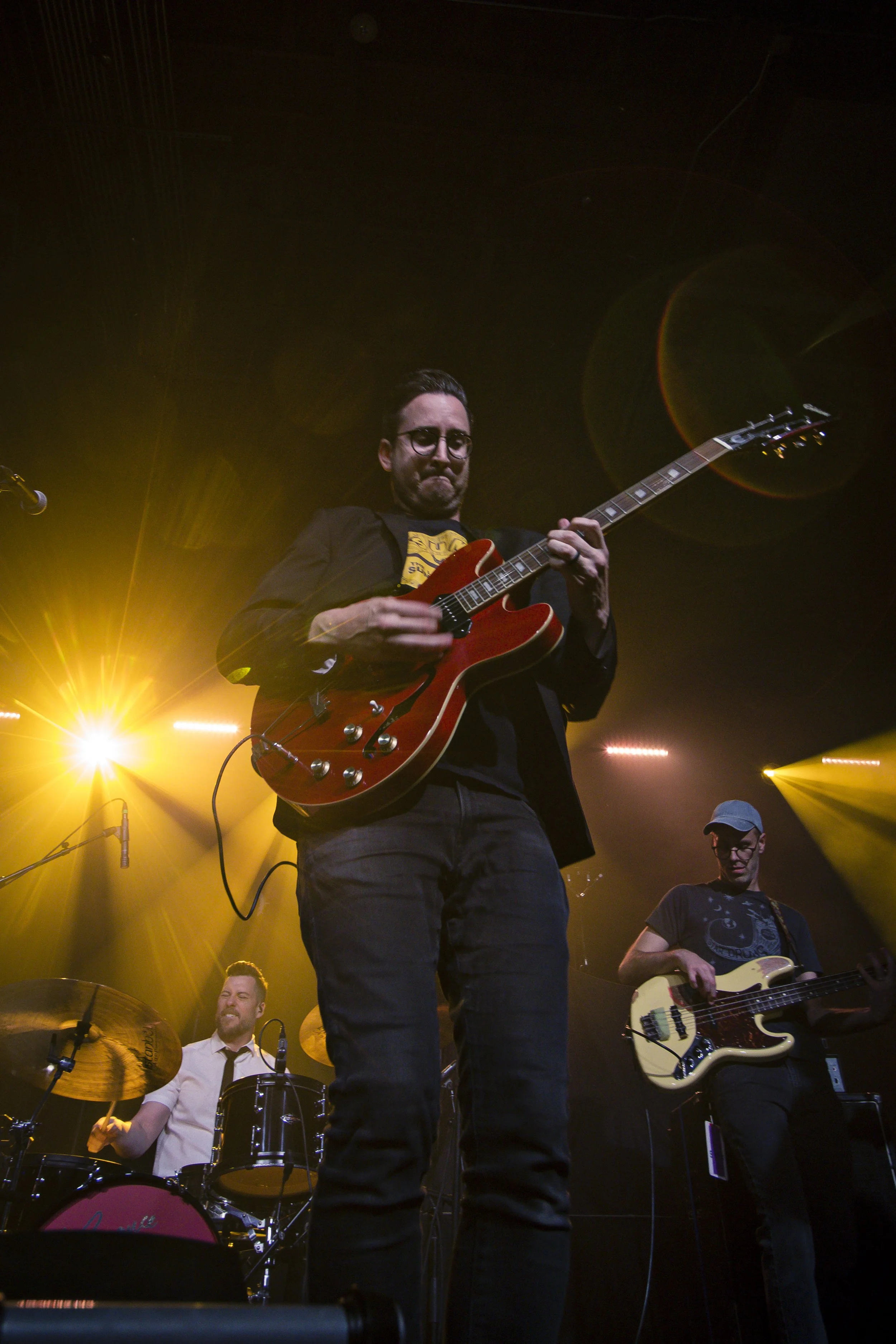 Musicians playing instruments on stage, with the lead guitarist in front, illuminated by yellow stage lights.