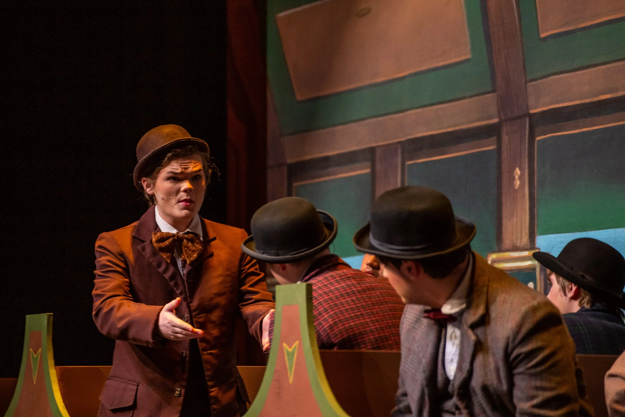 Actors in vintage costumes on stage, engaged in a theatrical performance, with one woman standing and speaking to the seated actors, all wearing bowler hats, in front of a painted backdrop of a building.