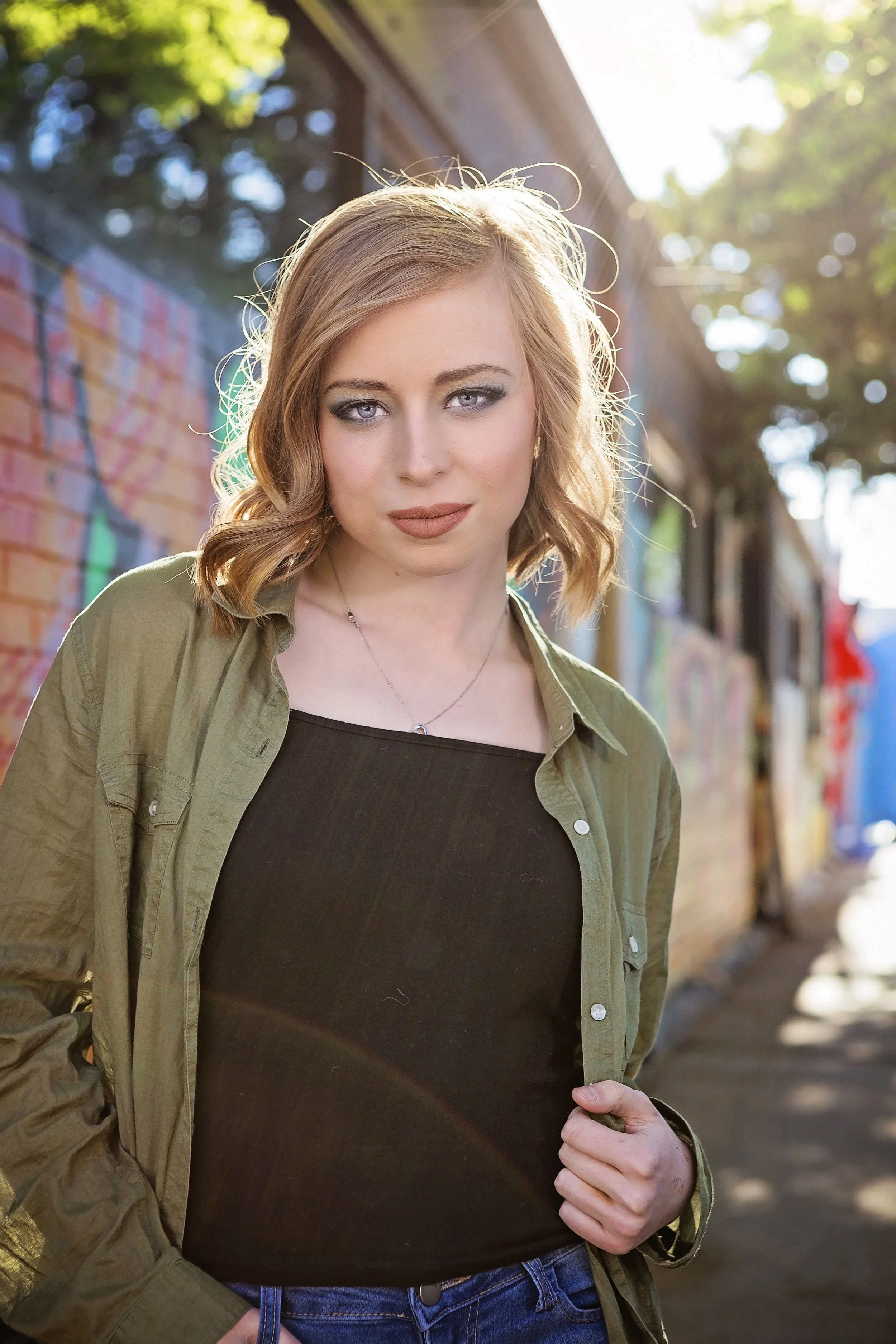 A young woman with shoulder-length wavy blonde hair and blue eyes stands outdoors in front of a colorful graffiti wall, with sunlight shining behind her. She wears a black top layered with an open green jacket and blue jeans.