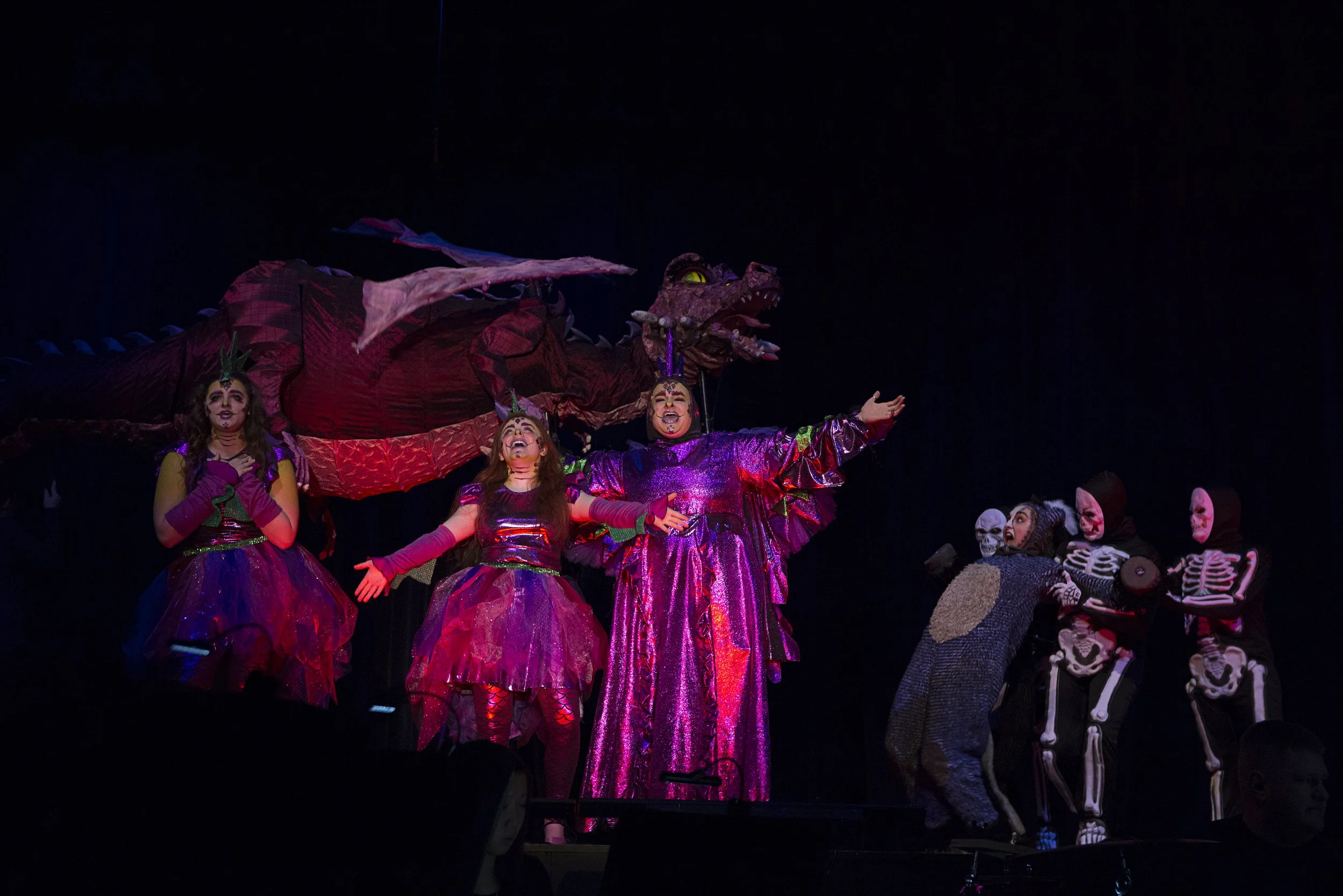 Theatrical performance with actors in colorful costumes, including a woman in a shiny purple dress, a girl in a pink dress, and a person with a dragon costume in the background, along with skeleton costumes, on a dark stage.